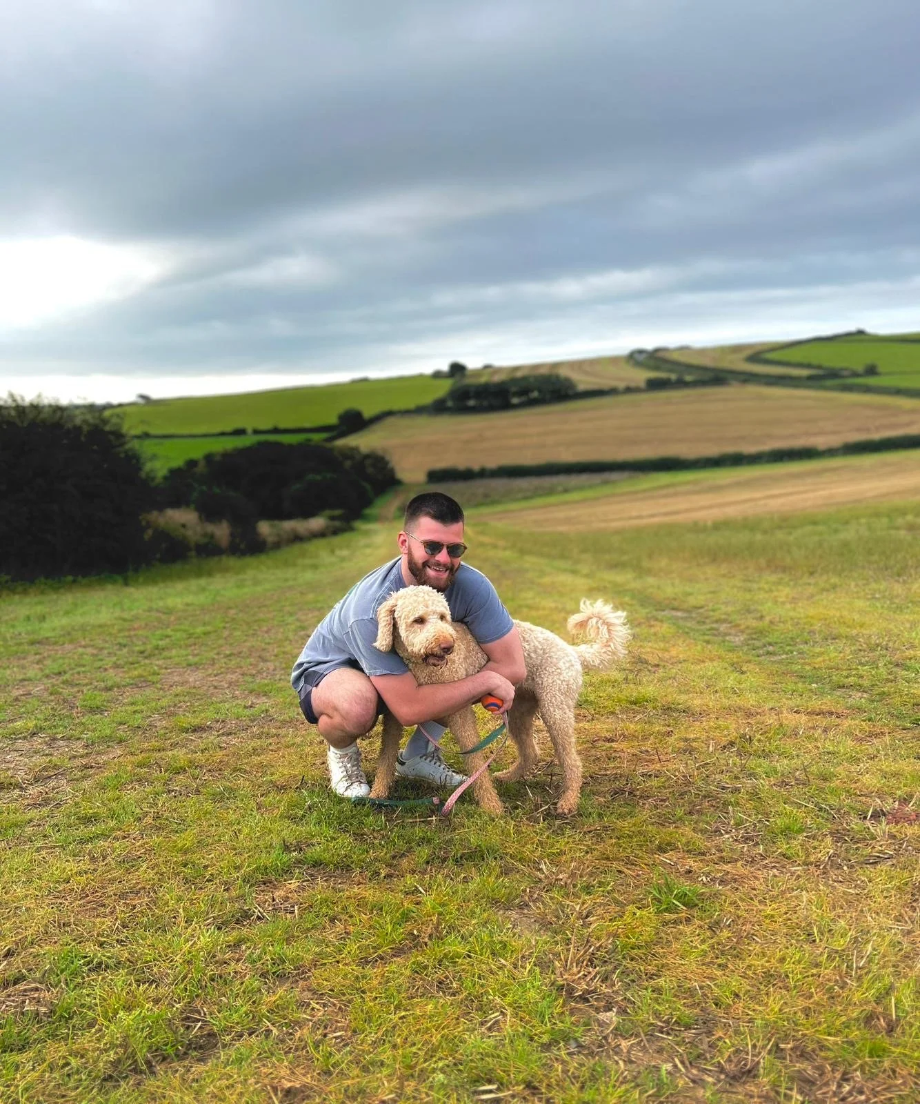 A man wearing sunglasses crouches down and hugs a large, curly-haired dog on a grassy field with rolling hills and a cloudy sky in the background.