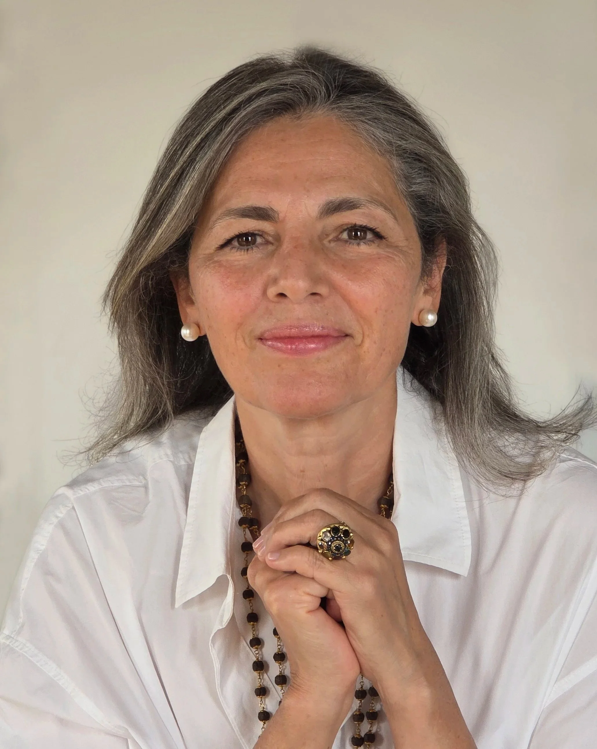 A middle-aged woman with shoulder-length gray hair, wearing a white shirt, pearl earrings, a gold ring, and a wood bead necklace, resting her chin on her clasped hands against a plain background.