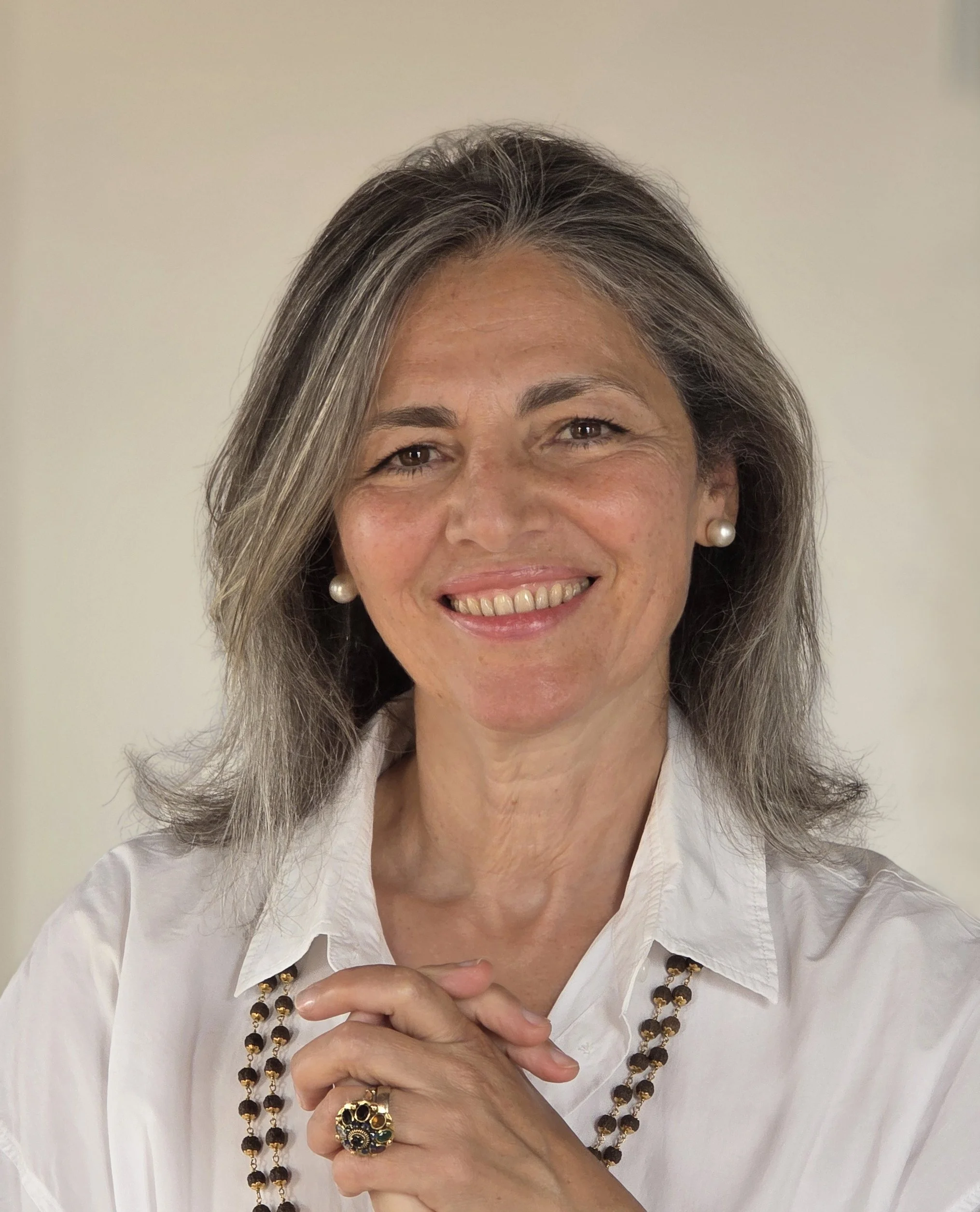 Close-up of a smiling woman with gray hair wearing a white shirt, pearl earrings, a necklace, and a large ring on her finger, holding her hands together in front of her.