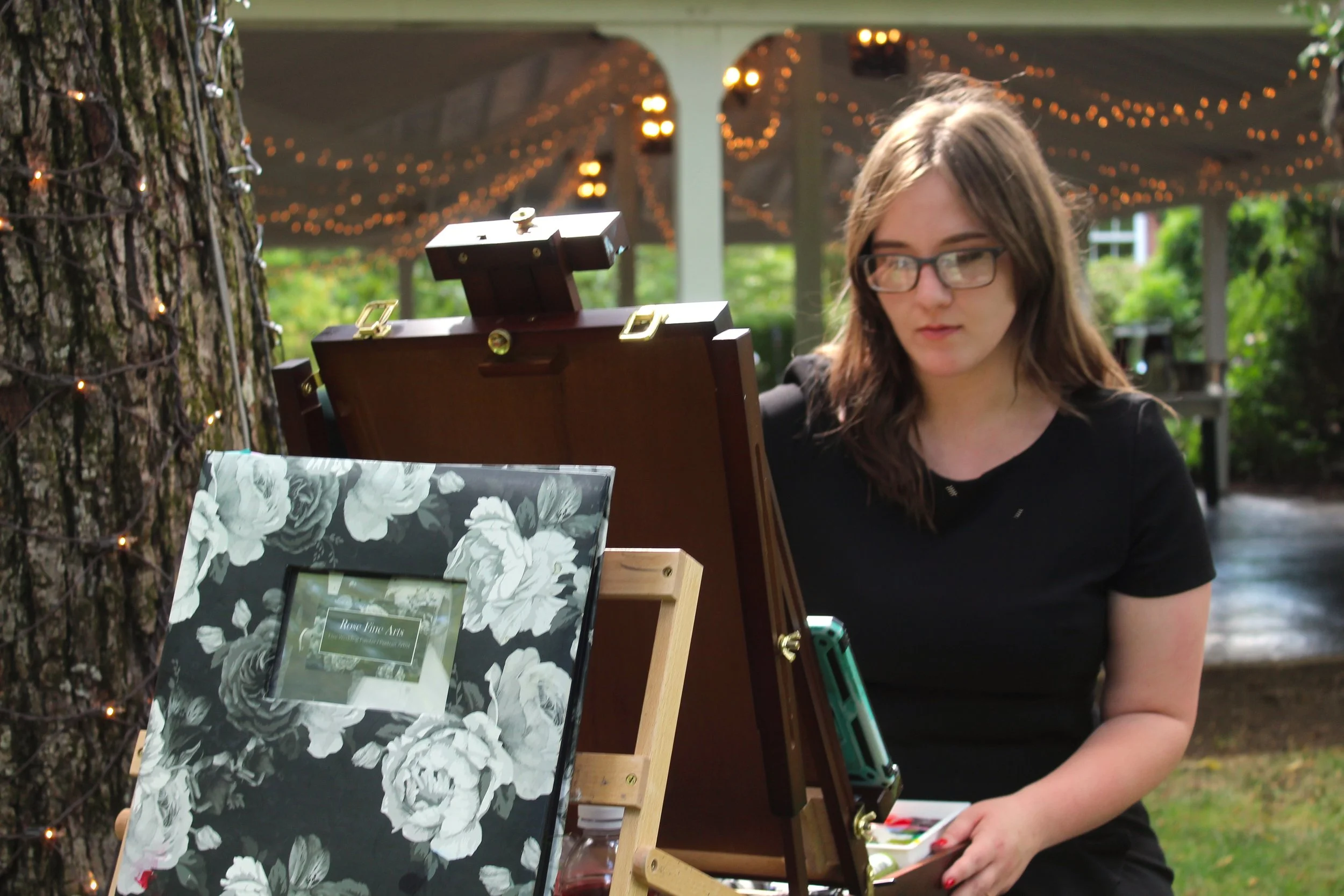 A young woman with glasses and long brown hair standing outdoors at night, looking at an easel with a painting or drawing, under string lights and a decorated pavilion.