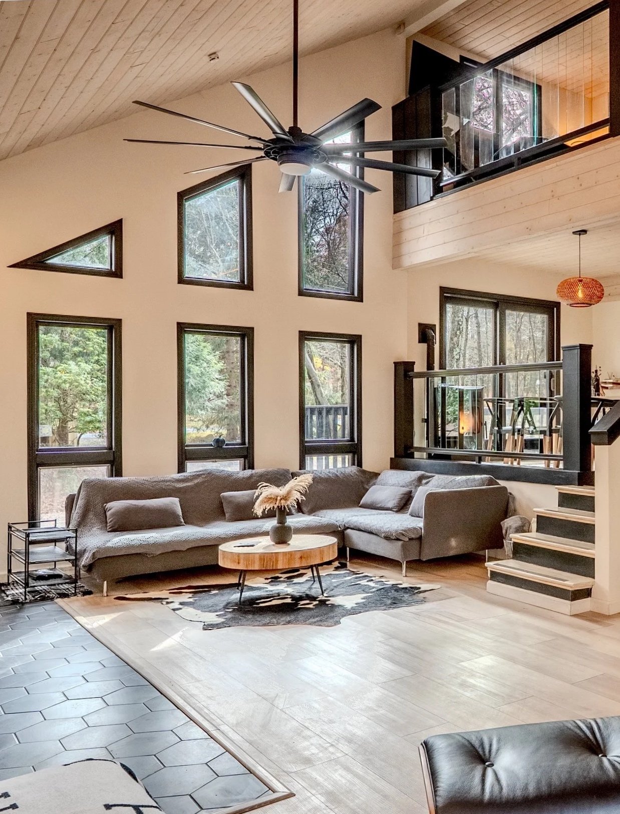 Living room with high ceiling, large windows, gray sectional sofa, round wooden coffee table, black and white cowhide rug, ceiling fan, stairs, and balcony in a modern home.