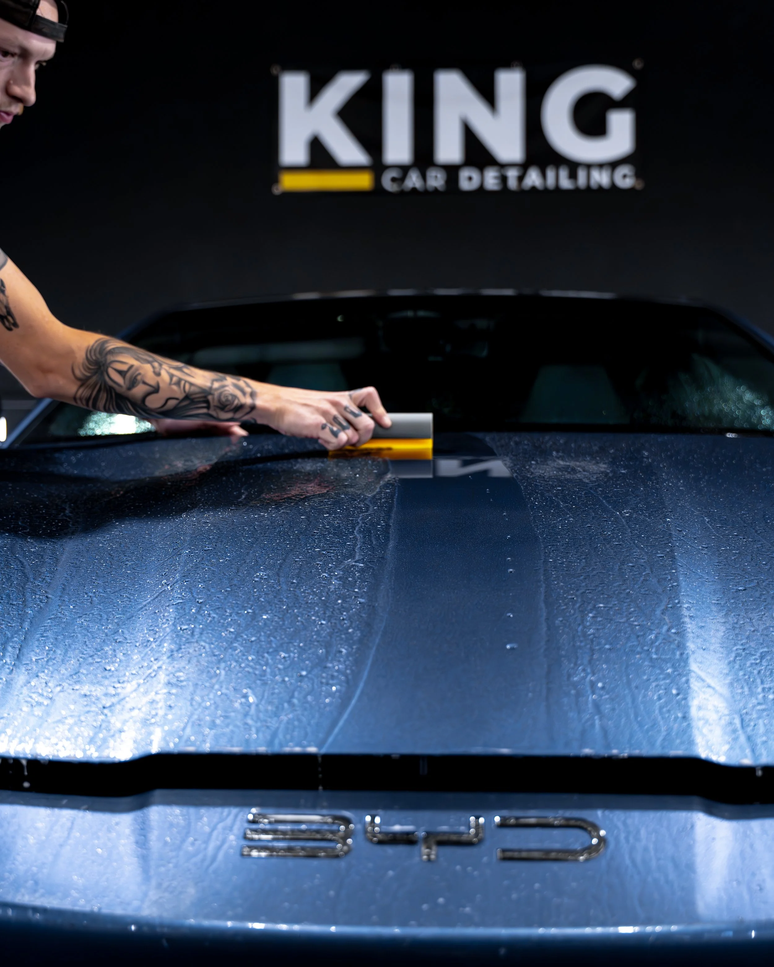 A person with tattoos is using a squeegee to apply ppf to the hood of a blue sports car at King Car Detailing, with water droplets visible on the surface.
