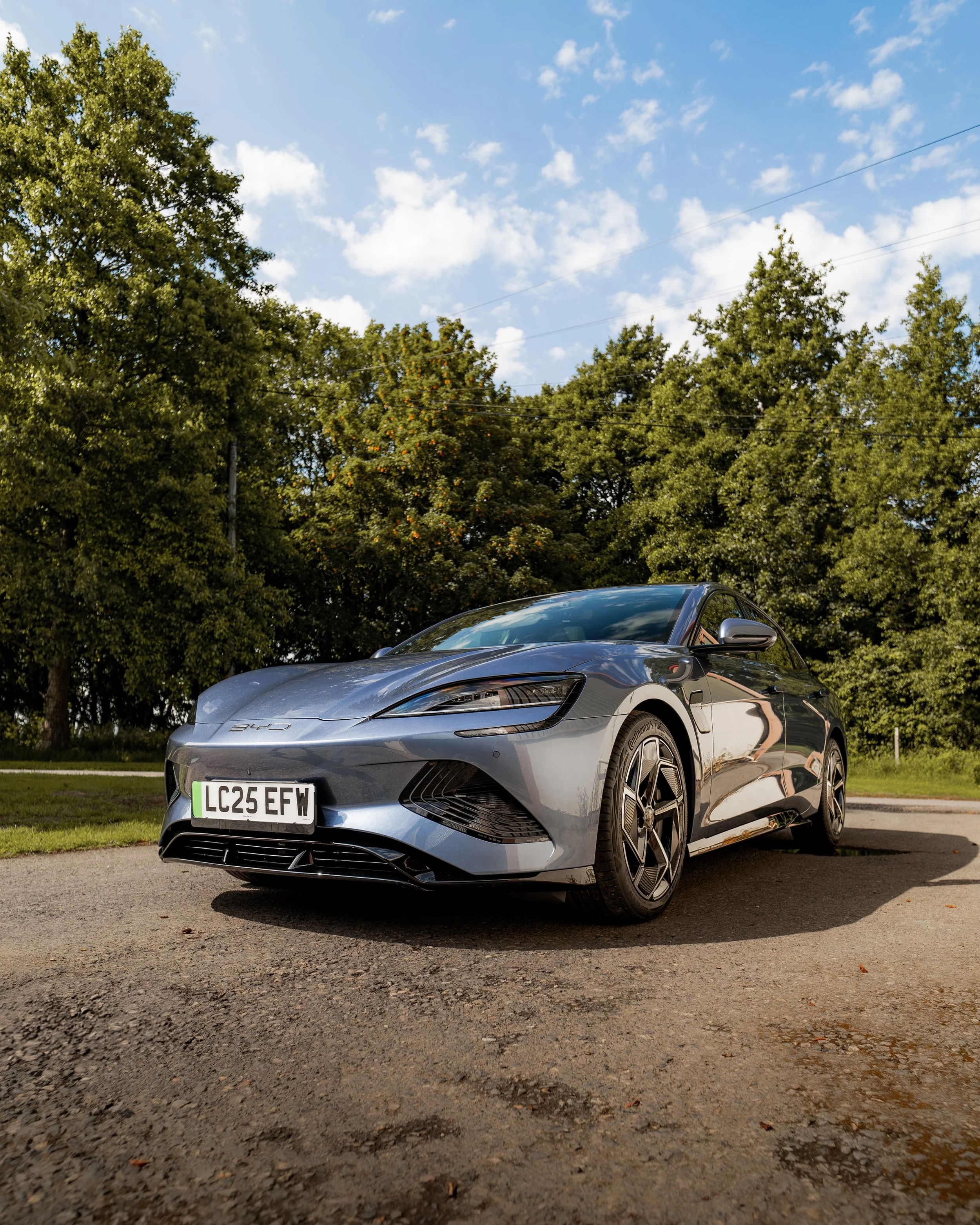 A silver sporty car parked on a gravel road with green trees and a blue sky in the background.