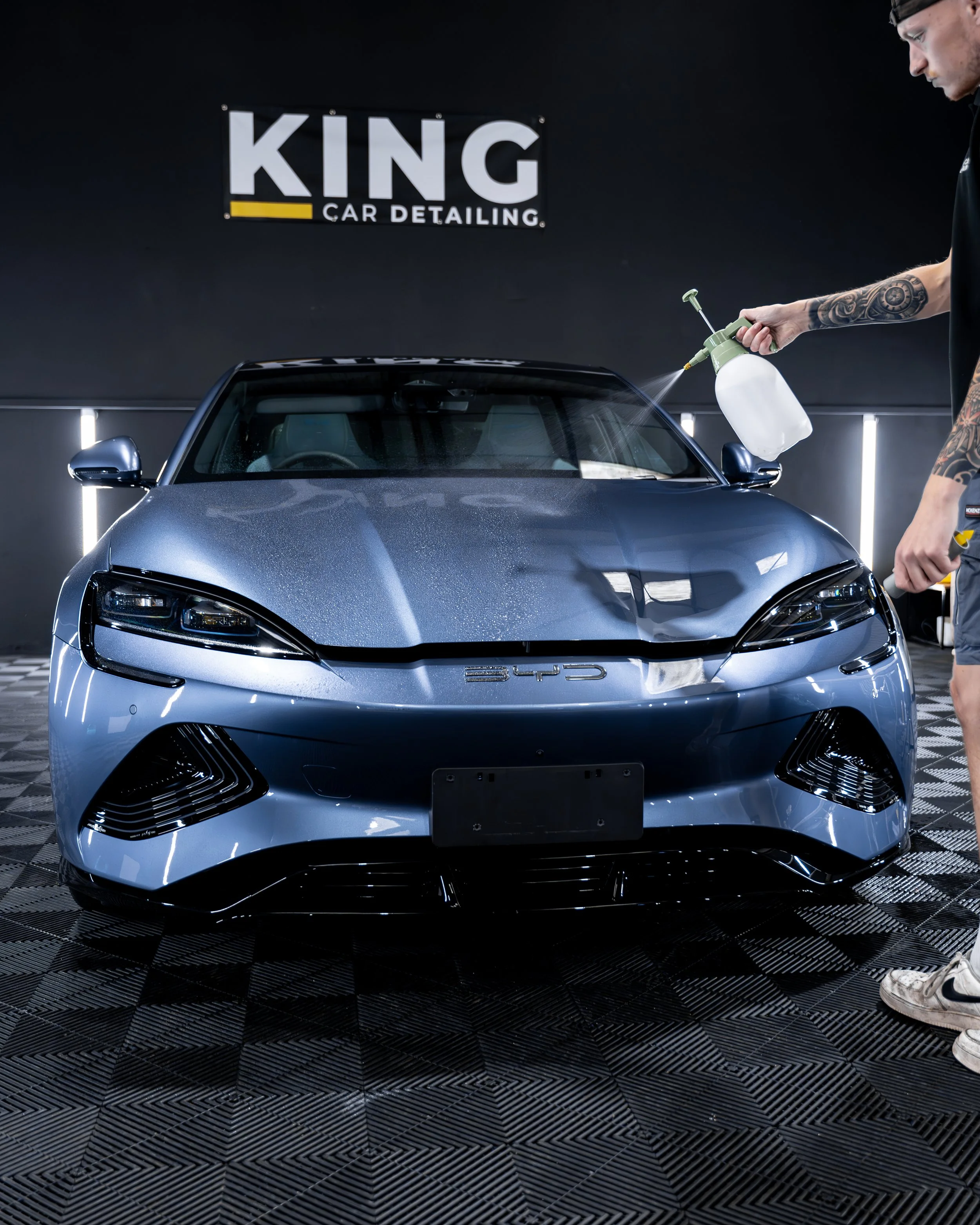 A person cleaning a blue electric car inside a detailing shop with a sign that reads 'KING CAR DETAILING' on the black wall behind them.
