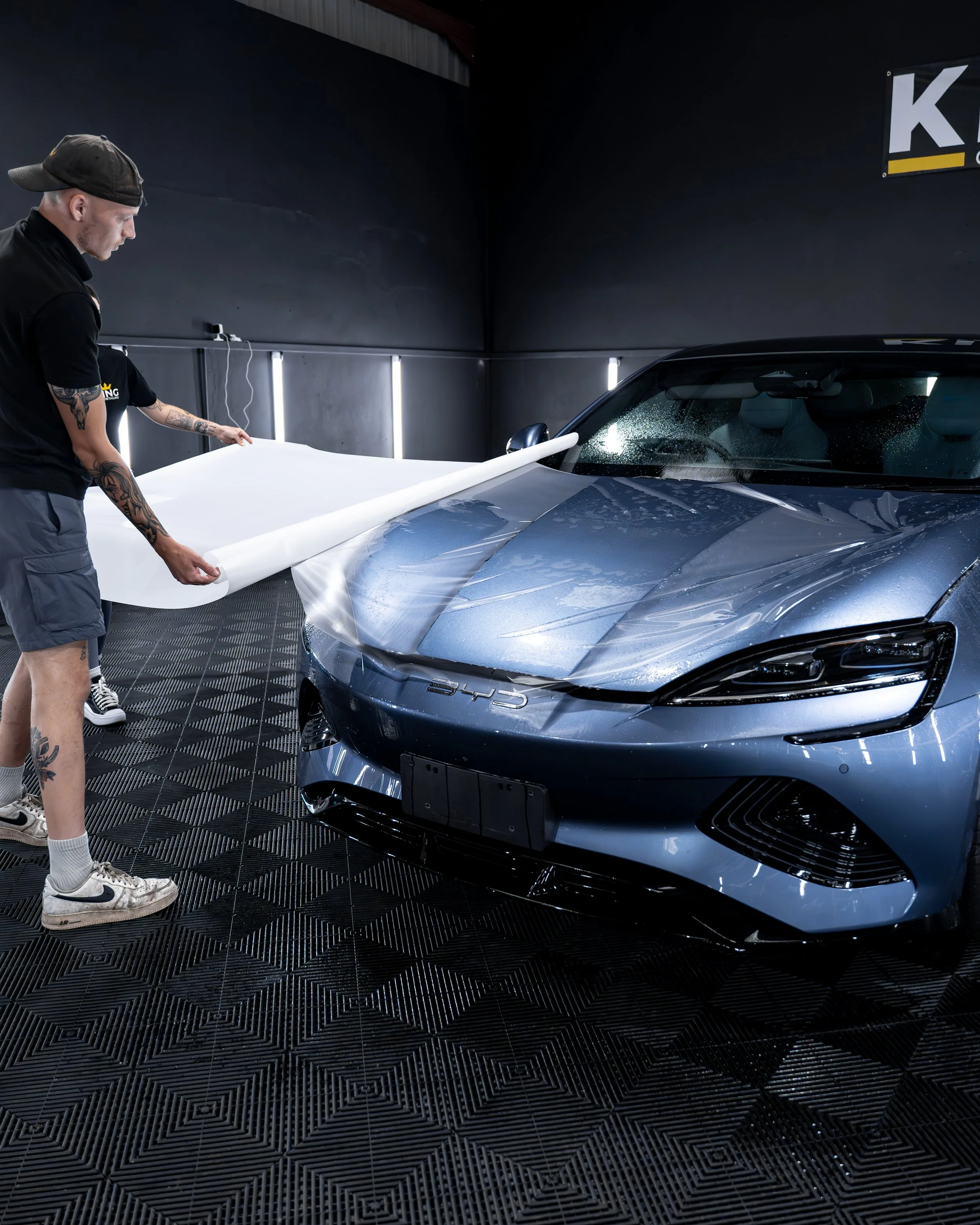 A man with tattoos and a cap is applying ppf to the hood of a blue sports car inside a workshop.