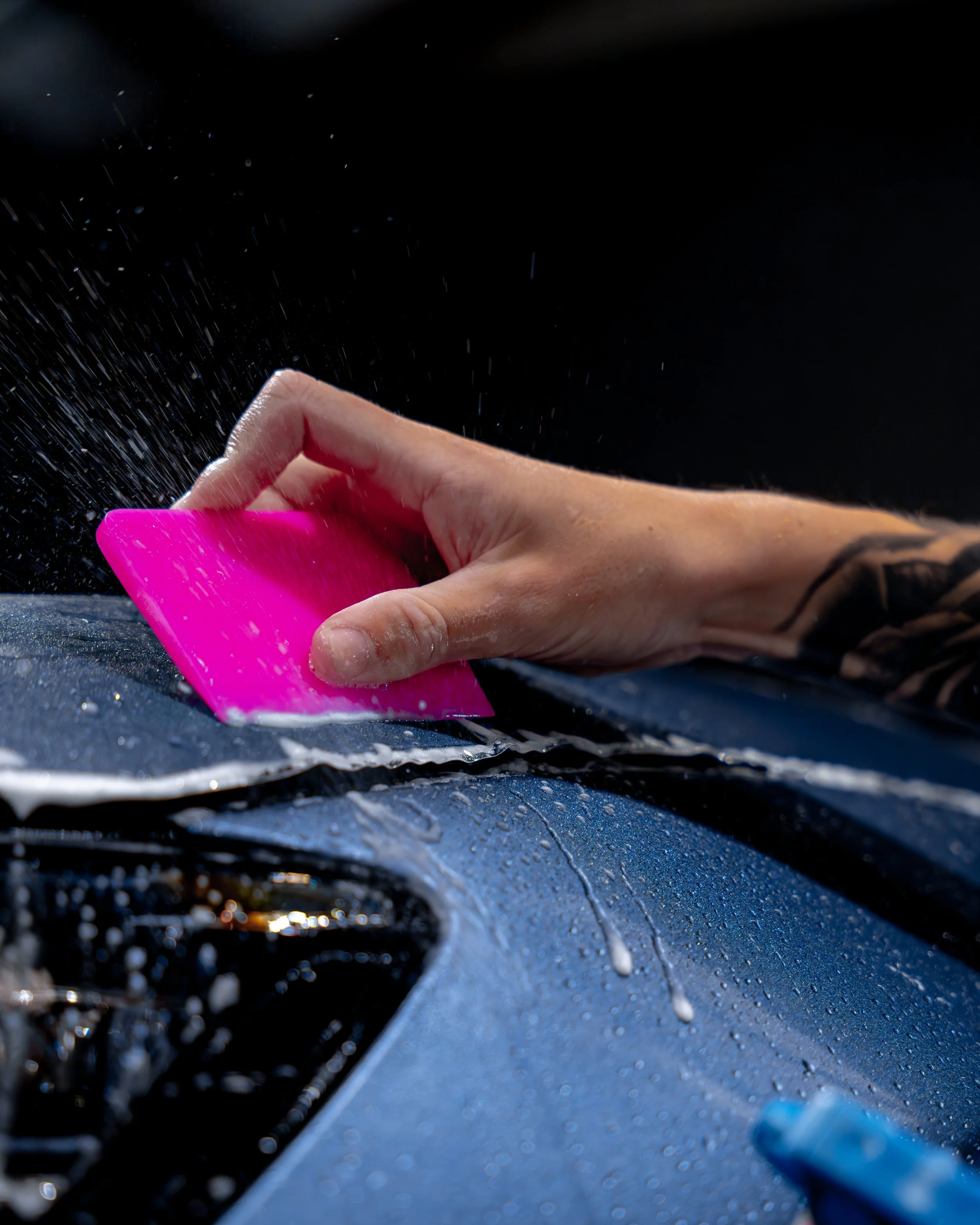 Hand using a pink sponge to wash the hood of a blue car, water and soap suds visible on the surface.