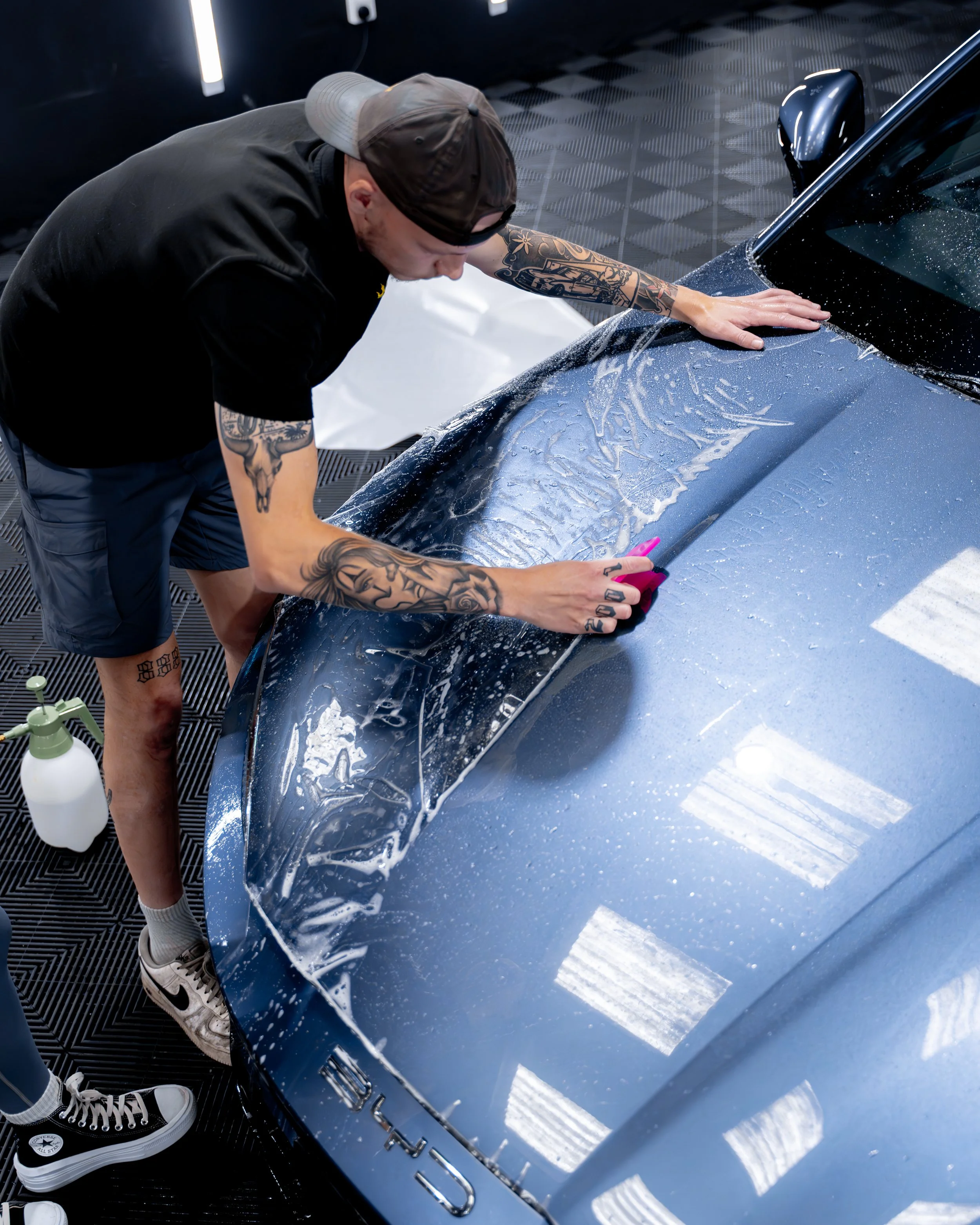 A man with tattoos, wearing a black T-shirt, shorts, and a backwards cap, is applying a protective film to a dark blue sports car inside a garage or detailing shop.