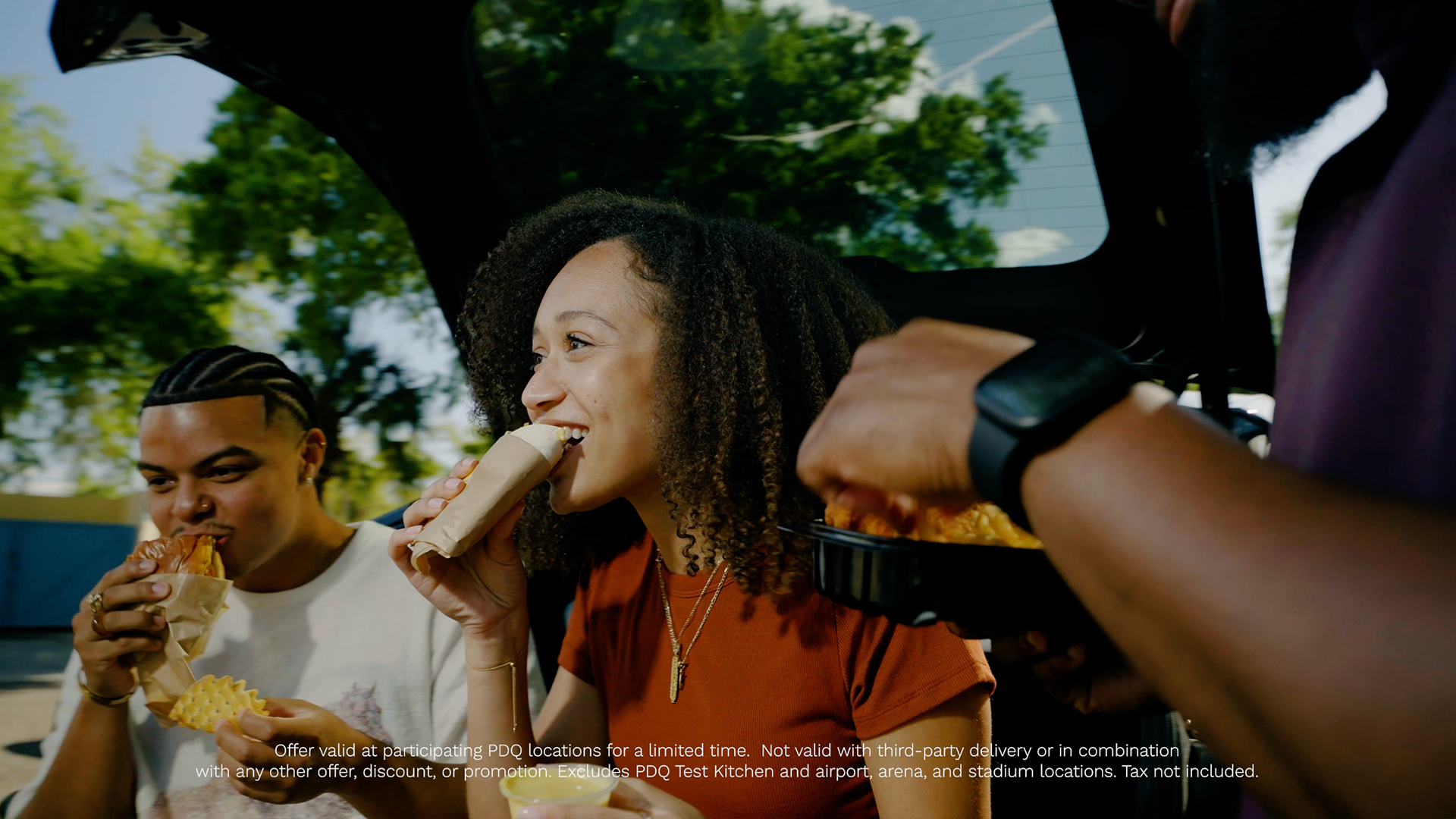 Two friends eating PDQ sandwiches and waffle fries together from the open trunk of a car