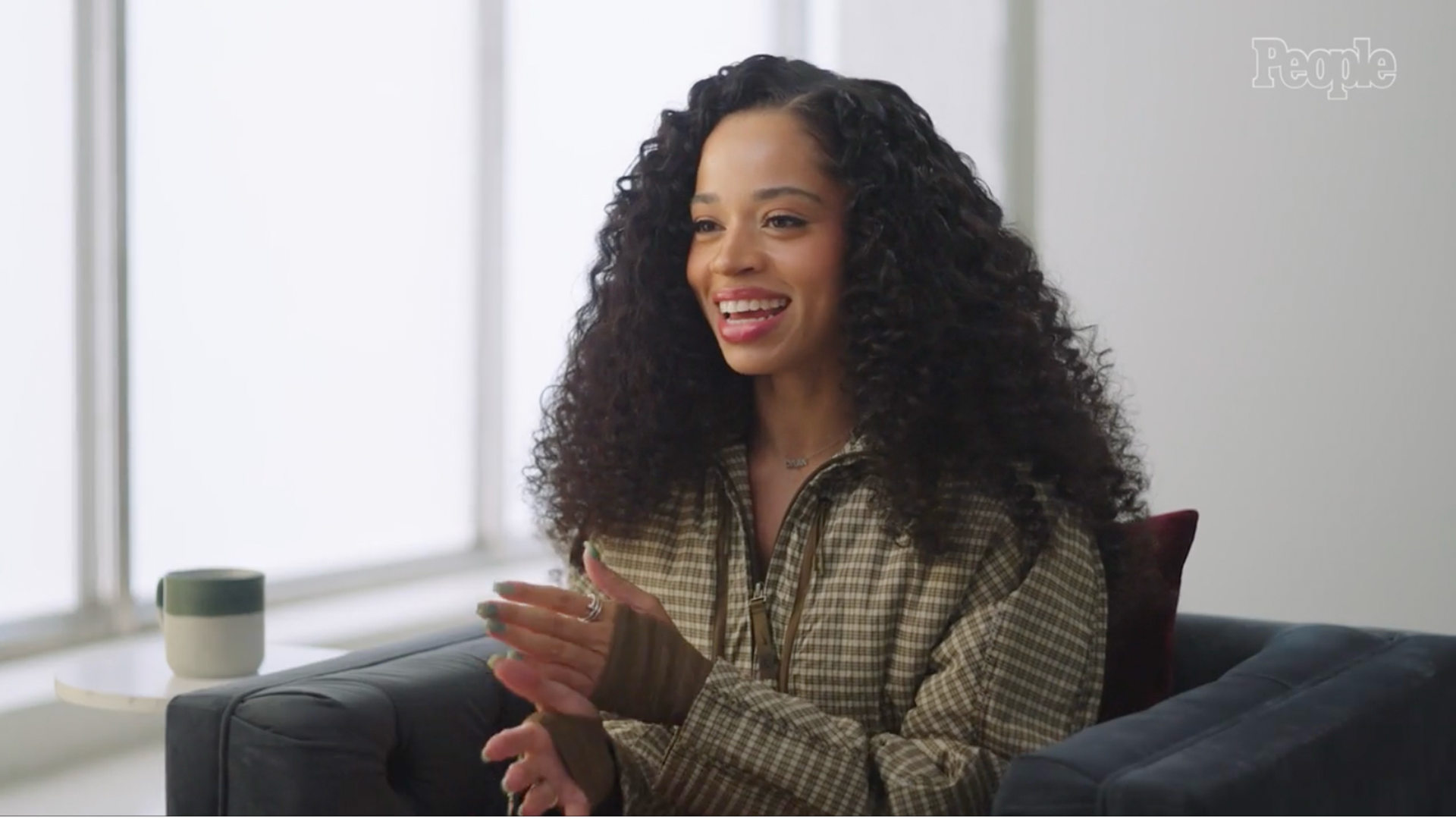 Ella Mai smiles and gestures while seated in a chair during an interview.