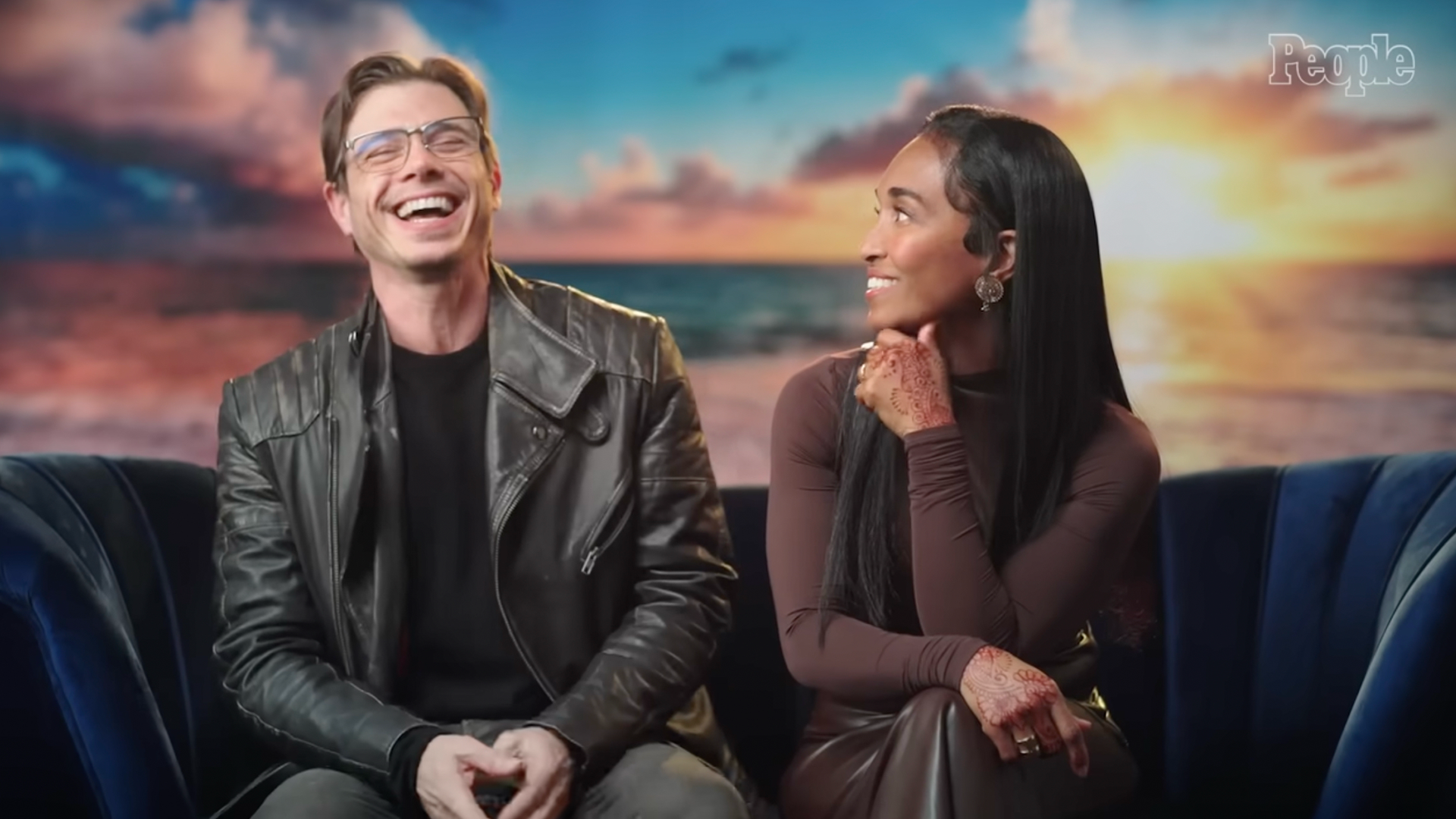 Matthew Lawrence laughs while sitting beside Chilli, who smiles at him, during an interview against a sunset backdrop