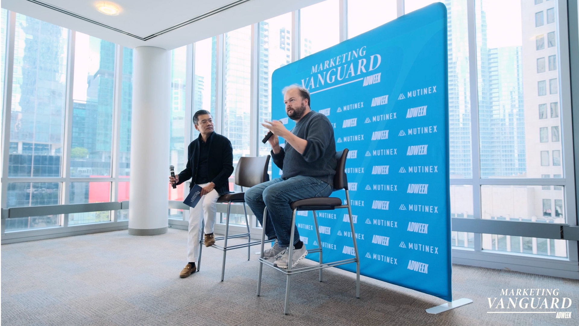 Two speakers holding microphones during an ADWEEK Marketing Vanguard panel in a bright event space overlooking the city