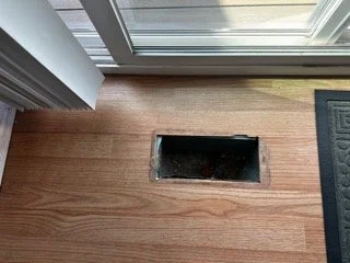 An open floor HVAC register vent cutout in wood-look laminate flooring used to inspect for hidden layers of older, suspect asbestos materials.