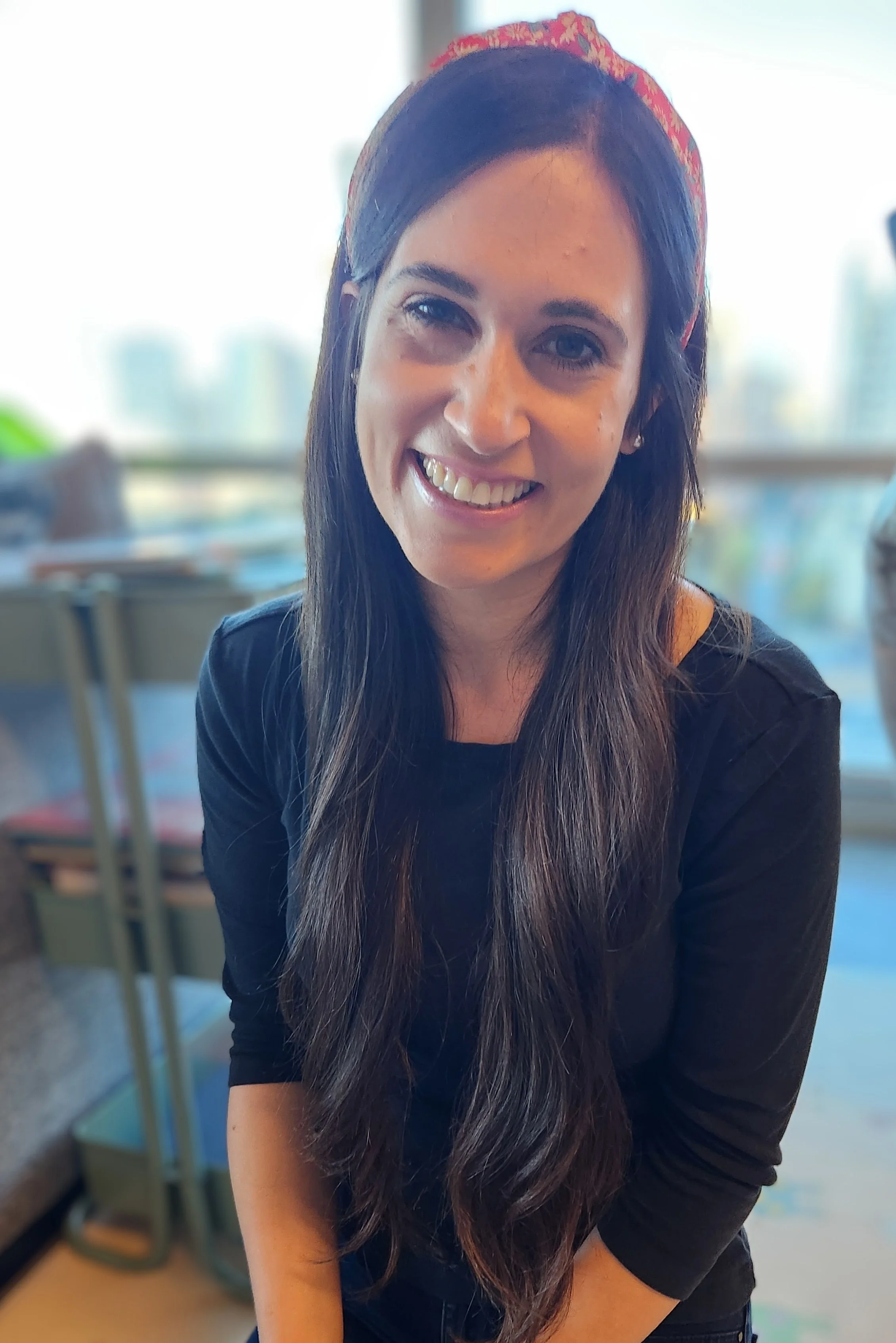 A woman with long brown hair, smiling, wearing a red headband and a black top, sitting indoors near a window with a cityscape in the background.