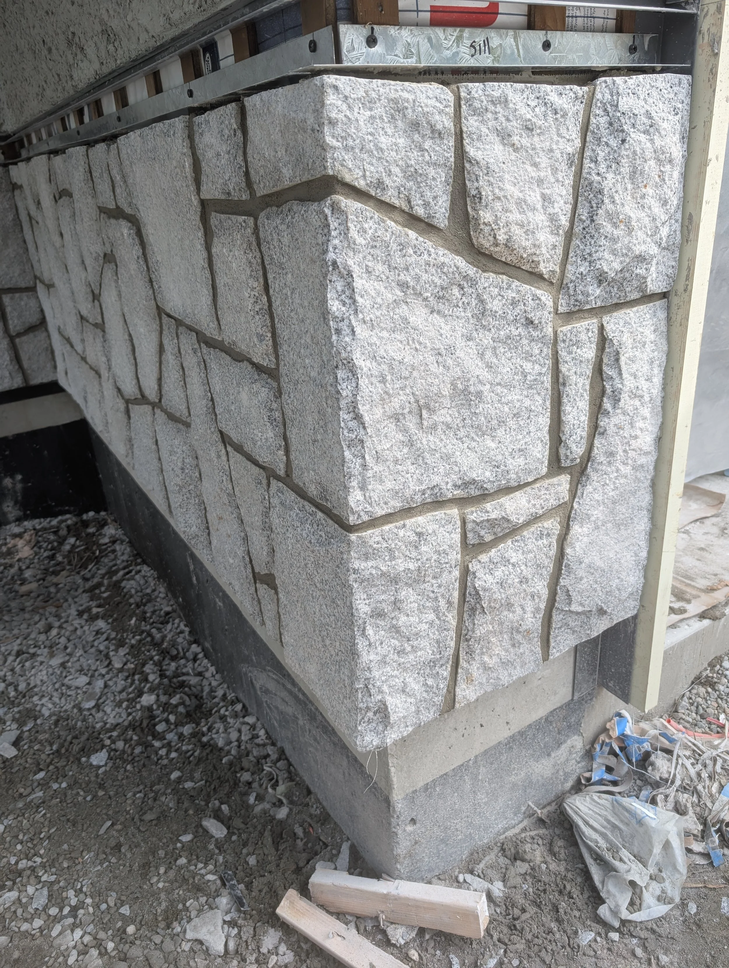 Close-up view of a stone veneer wall under construction, with mortar and tools visible, and gravel on the ground.