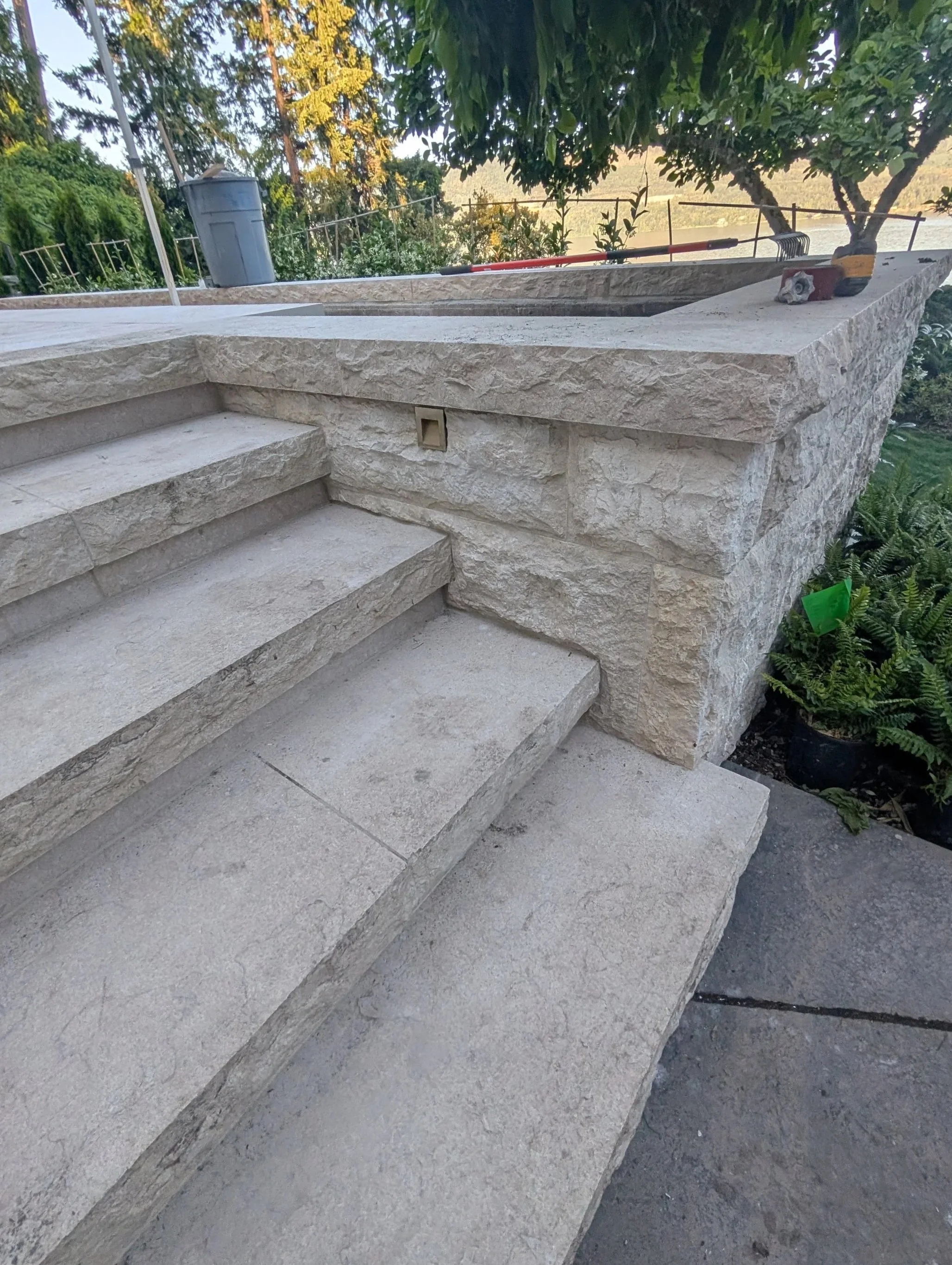 Stone outdoor staircase with four steps, leading to a flat surface with stone walls on either side. Green foliage and trees in the background, along with a black metal railing, a trash bin, and some gardening tools. Overcast sky, natural outdoor sett