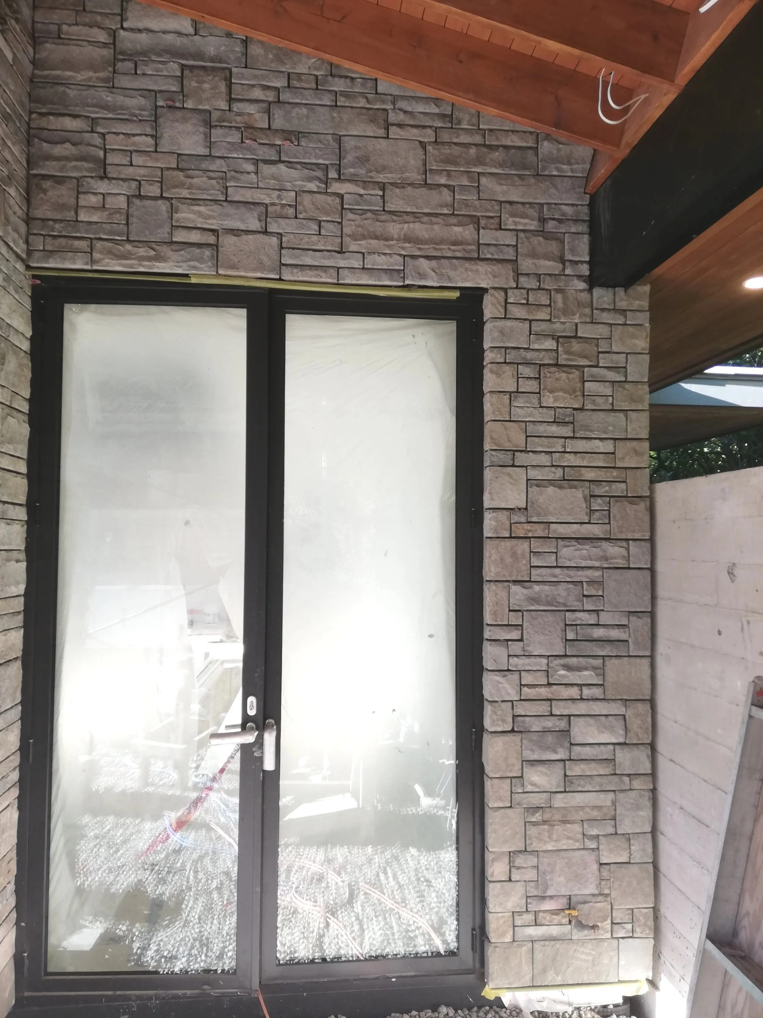 Exterior of a building with a black framed glass door, stone and brick wall, and a wooden ceiling.