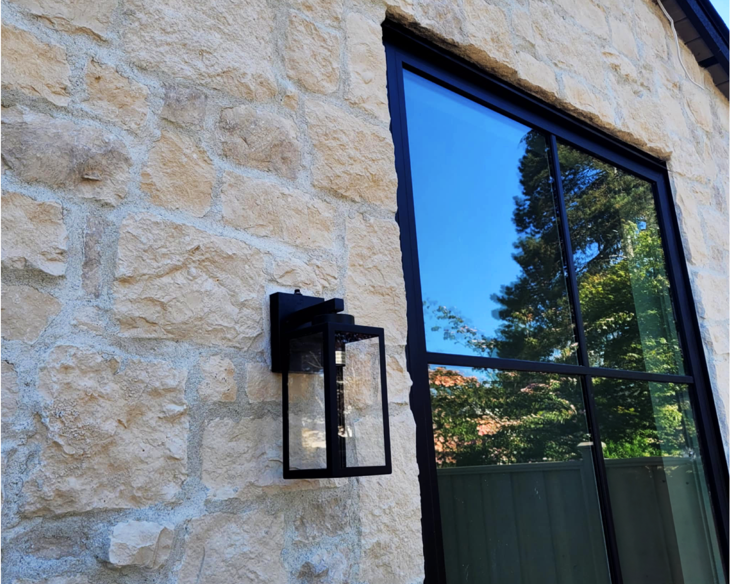 Close-up of a stone exterior wall with a large window reflecting trees and blue sky. Sandstone masonry design on a residential home in north vancouver