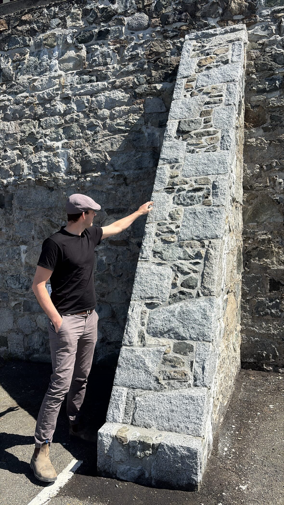 A man in a black shirt, beige pants, and beige boots is standing outdoors next to a large stone monument or sculpture made of stacked rough-cut stones. The man is touching the sculpture with his left hand and looking at it.