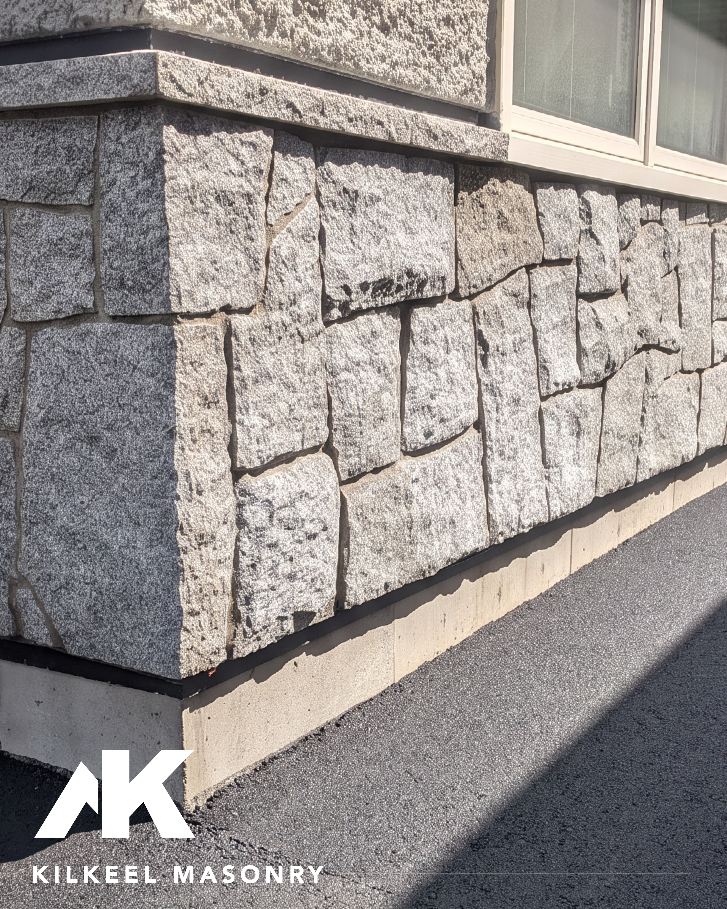 Close-up of a stone building corner with textured gray stone bricks and a window with white trim; at the bottom, white text reads 'KILKEEL MASONRY' with part of a logo visible.