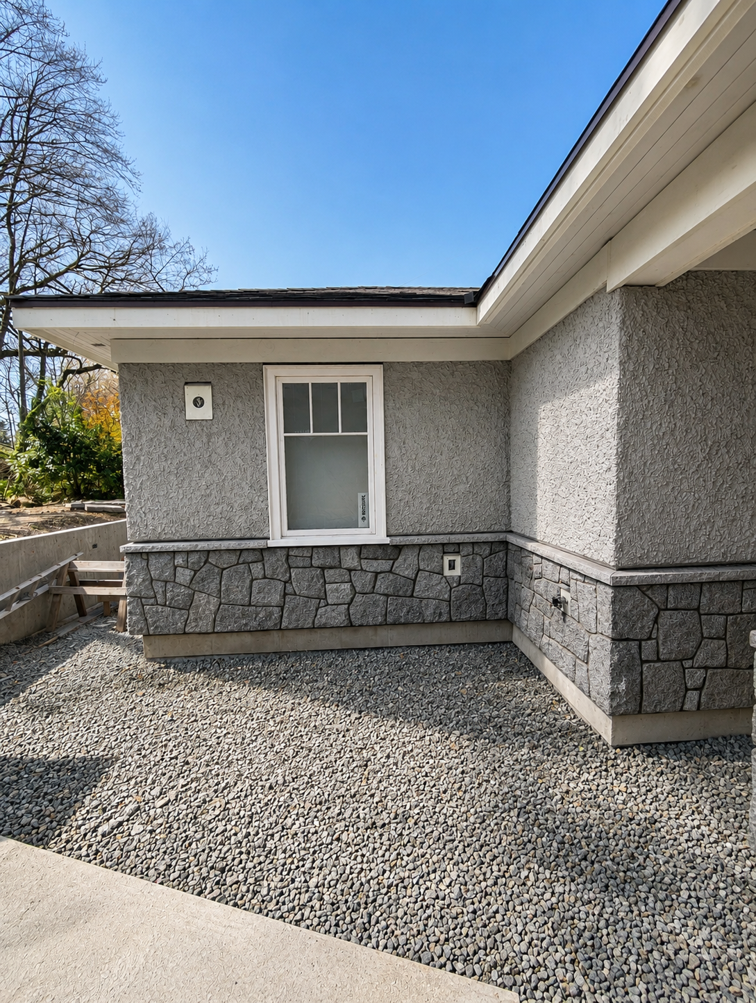 Exterior corner of a house with gray textured stucco and stone veneer, white window, gravel ground, and a blue sky.