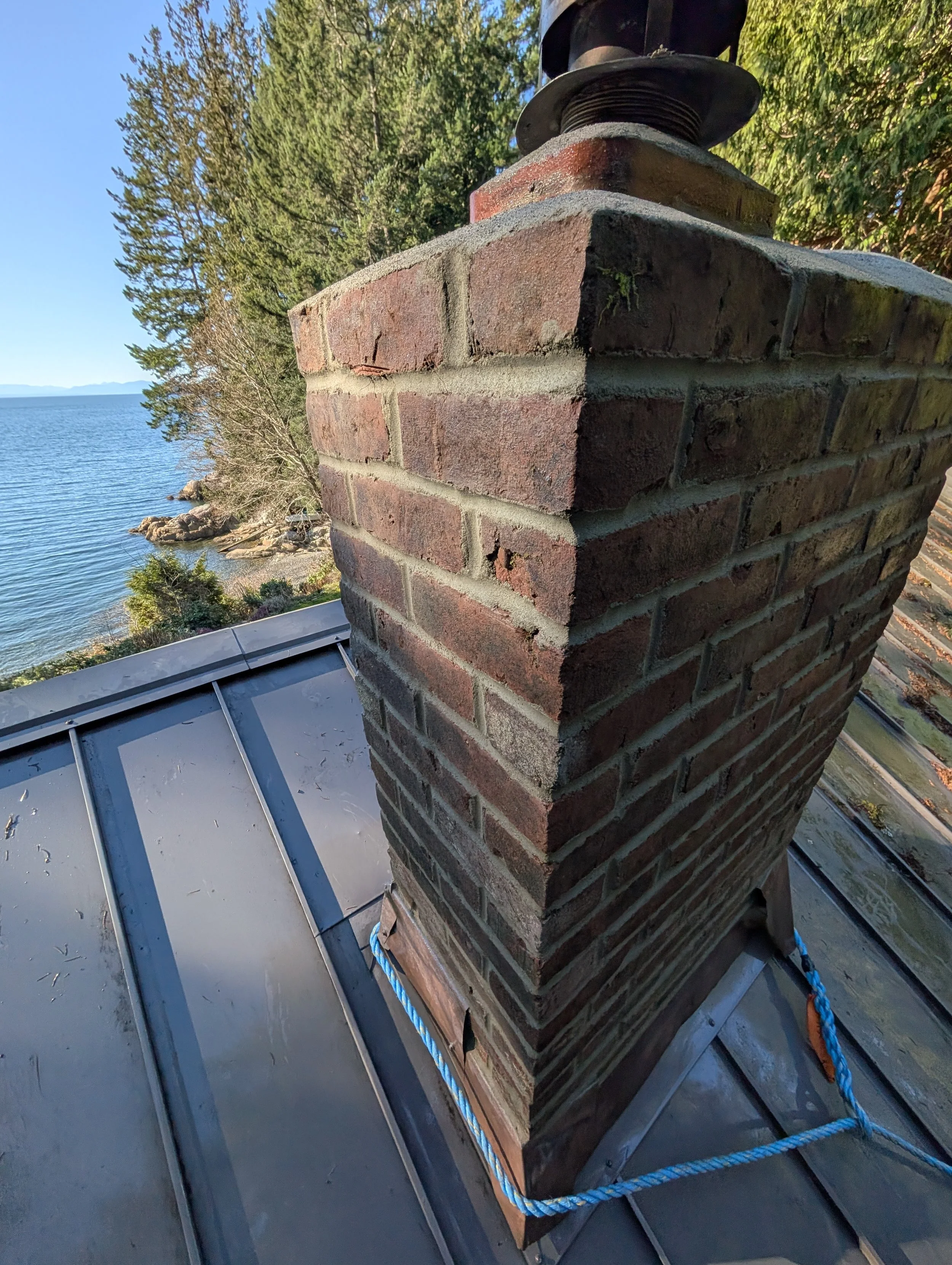 Close-up of a brick chimney on a metal roof near a lakeside, with trees and water in the background on a clear, sunny day. Chimney restoration on bowen island