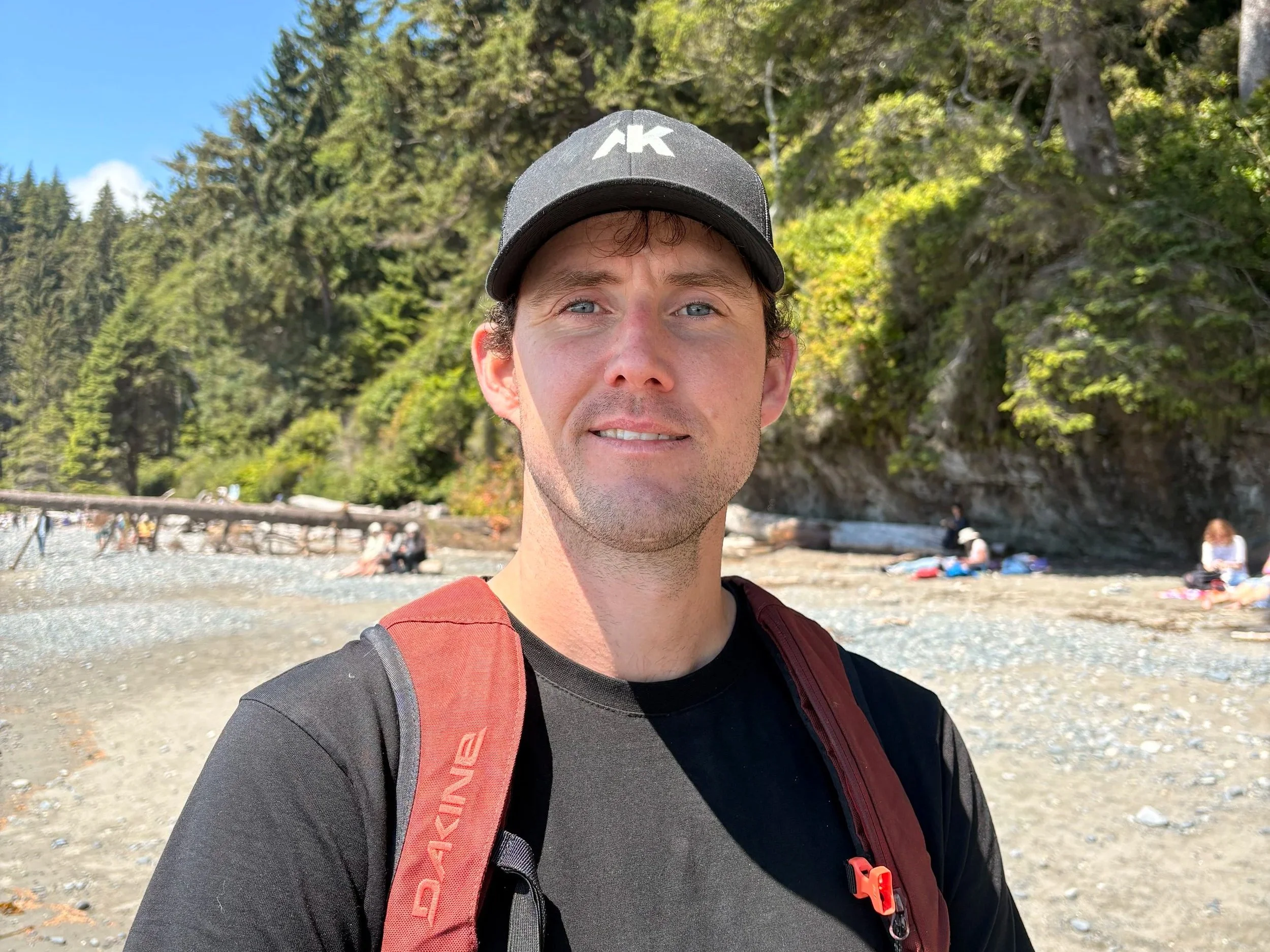 A man with curly brown hair, blue eyes, and a slight smile, wearing a gray baseball cap with a white logo, a black shirt, and a red backpack, standing outdoors with a forested background and a beach with people.
