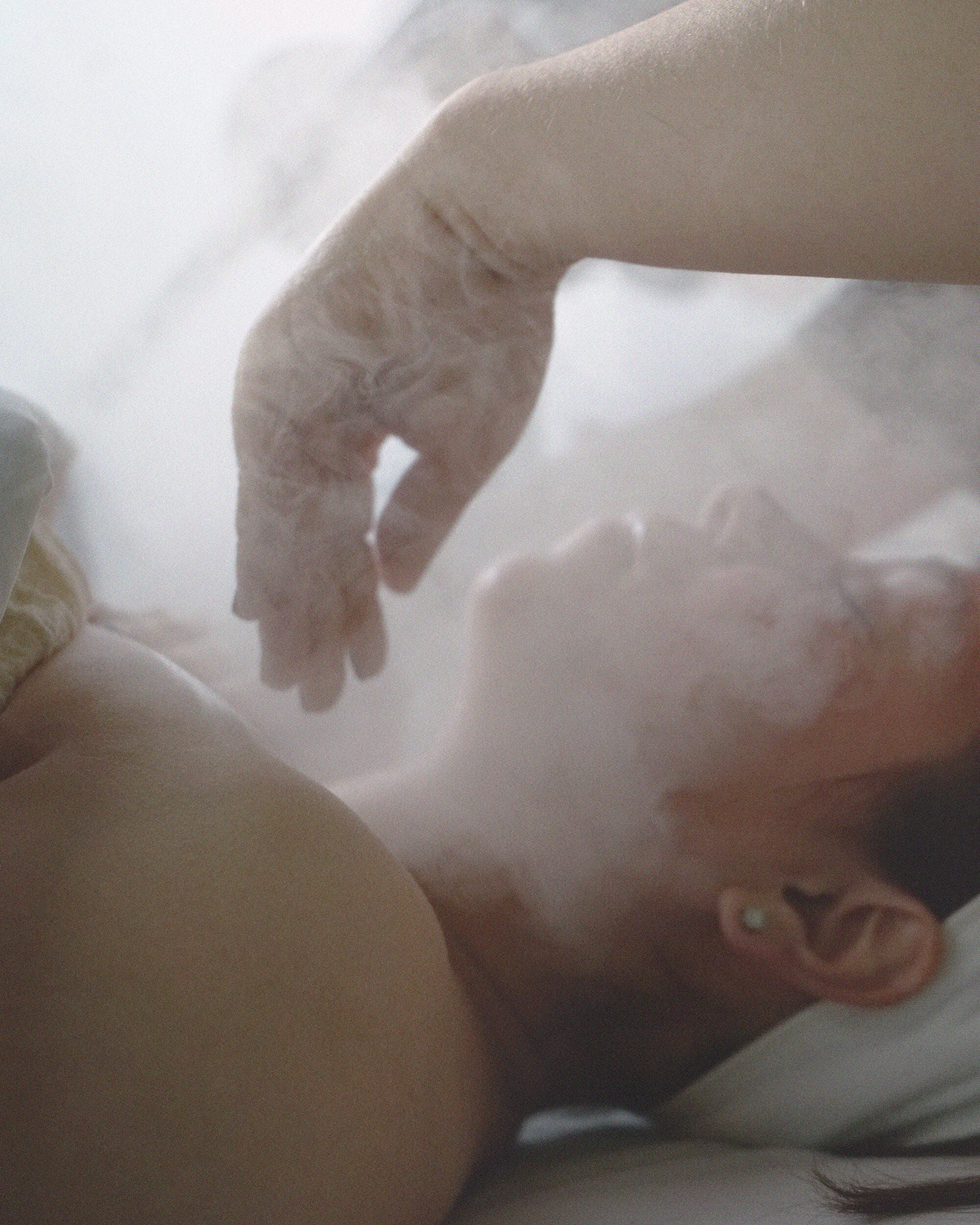 A woman lying on a treatment table with eyes closed, receiving a facial with steam. The scene is soft and intimate.