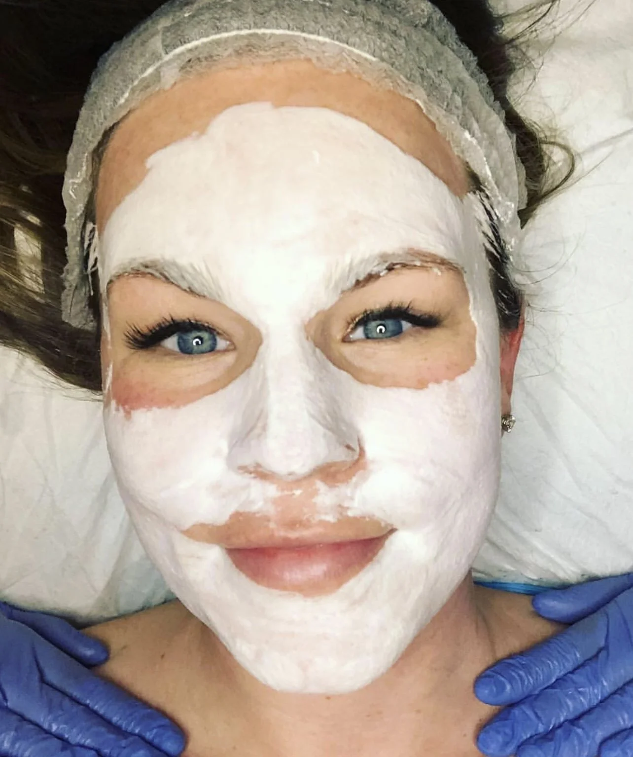 A woman with blue eyes lying on a bed with a white face mask applied to her face, wearing a headband and blue gloves.