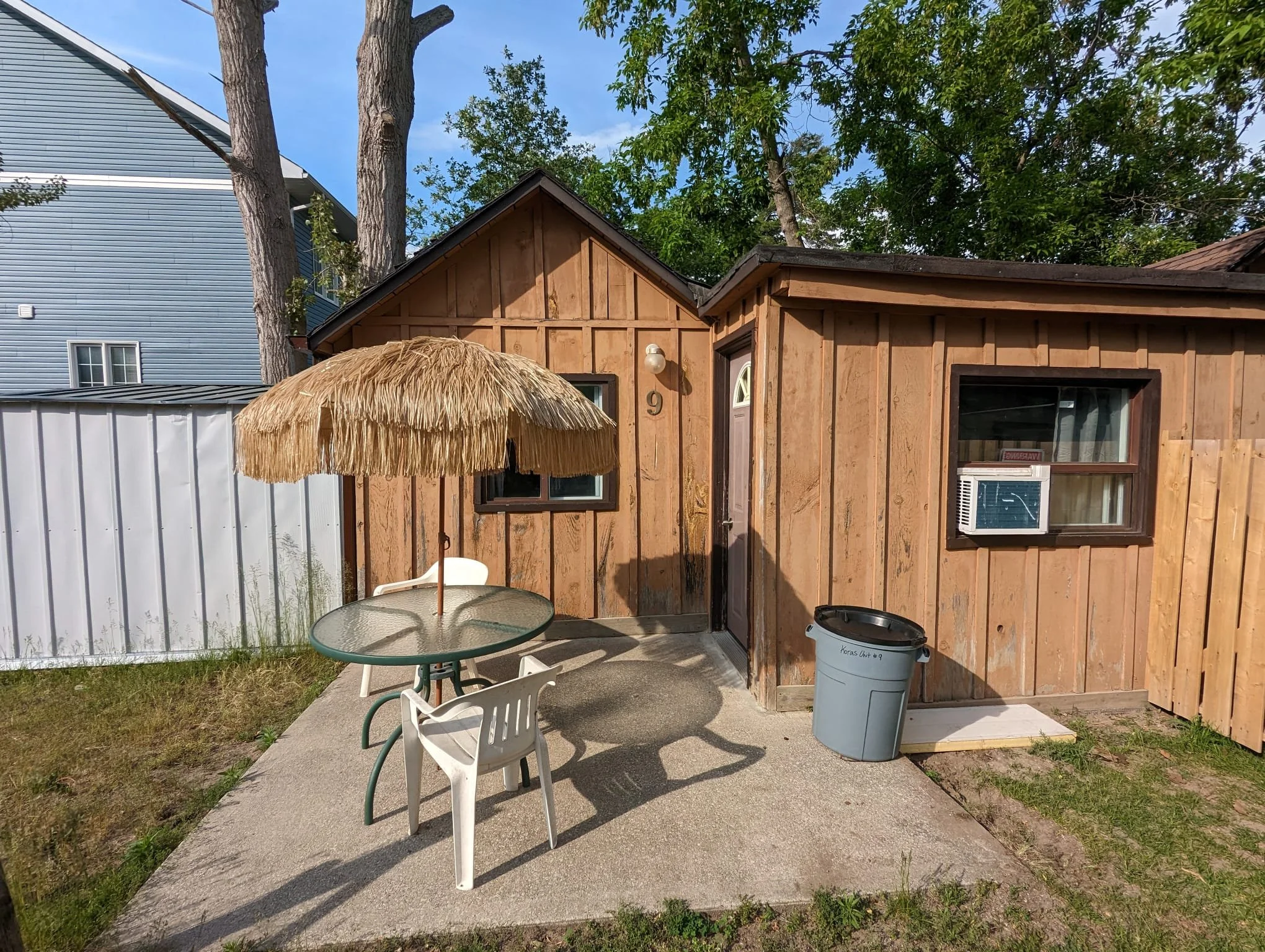 Backyard patio with a round glass table, white plastic chairs, a thatched umbrella, and a small wooden shed with a window air conditioning unit.