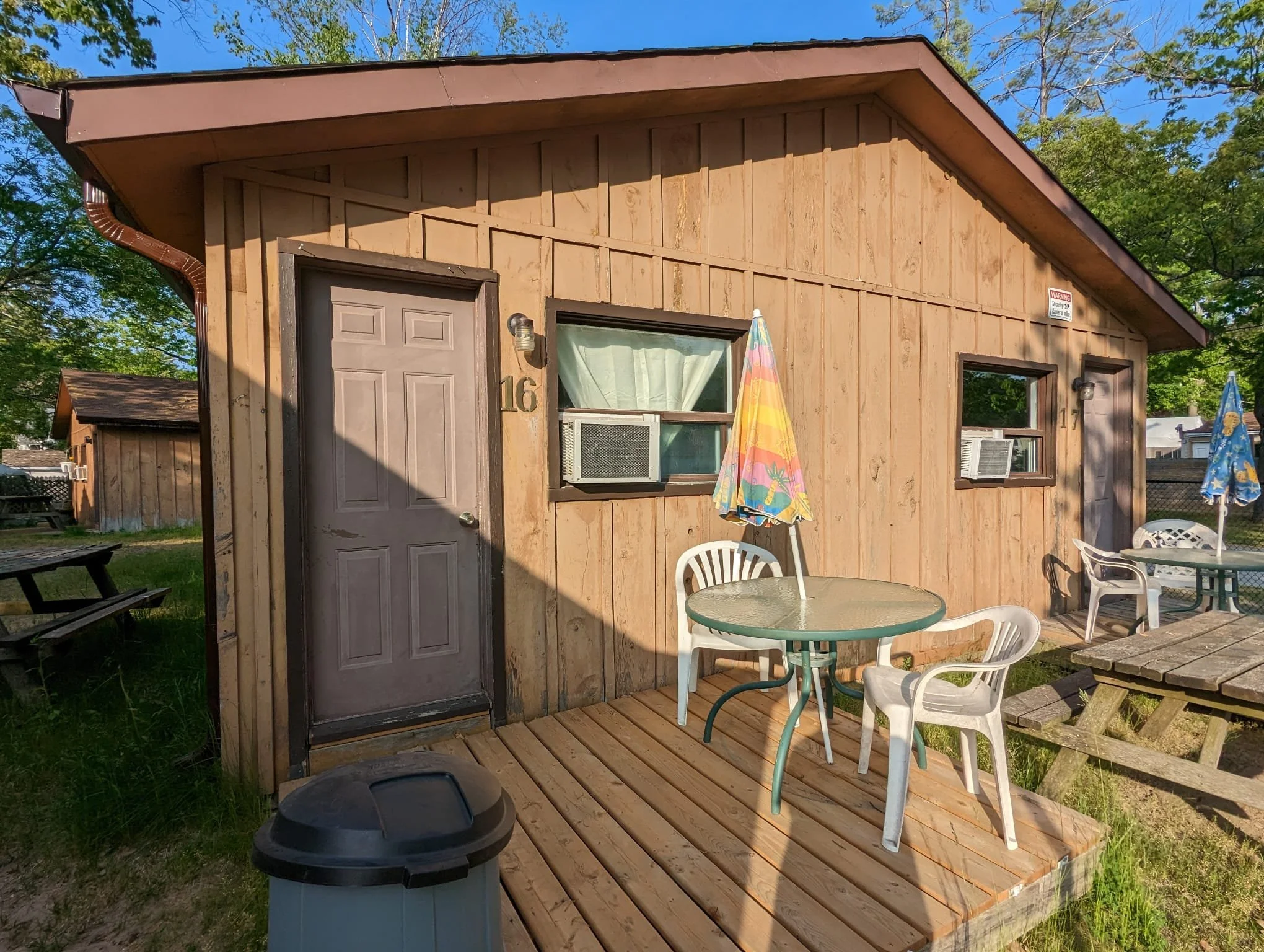 Small wooden cabin with a deck, outdoor table, chairs, and umbrellas, in a yard with grass and trees.