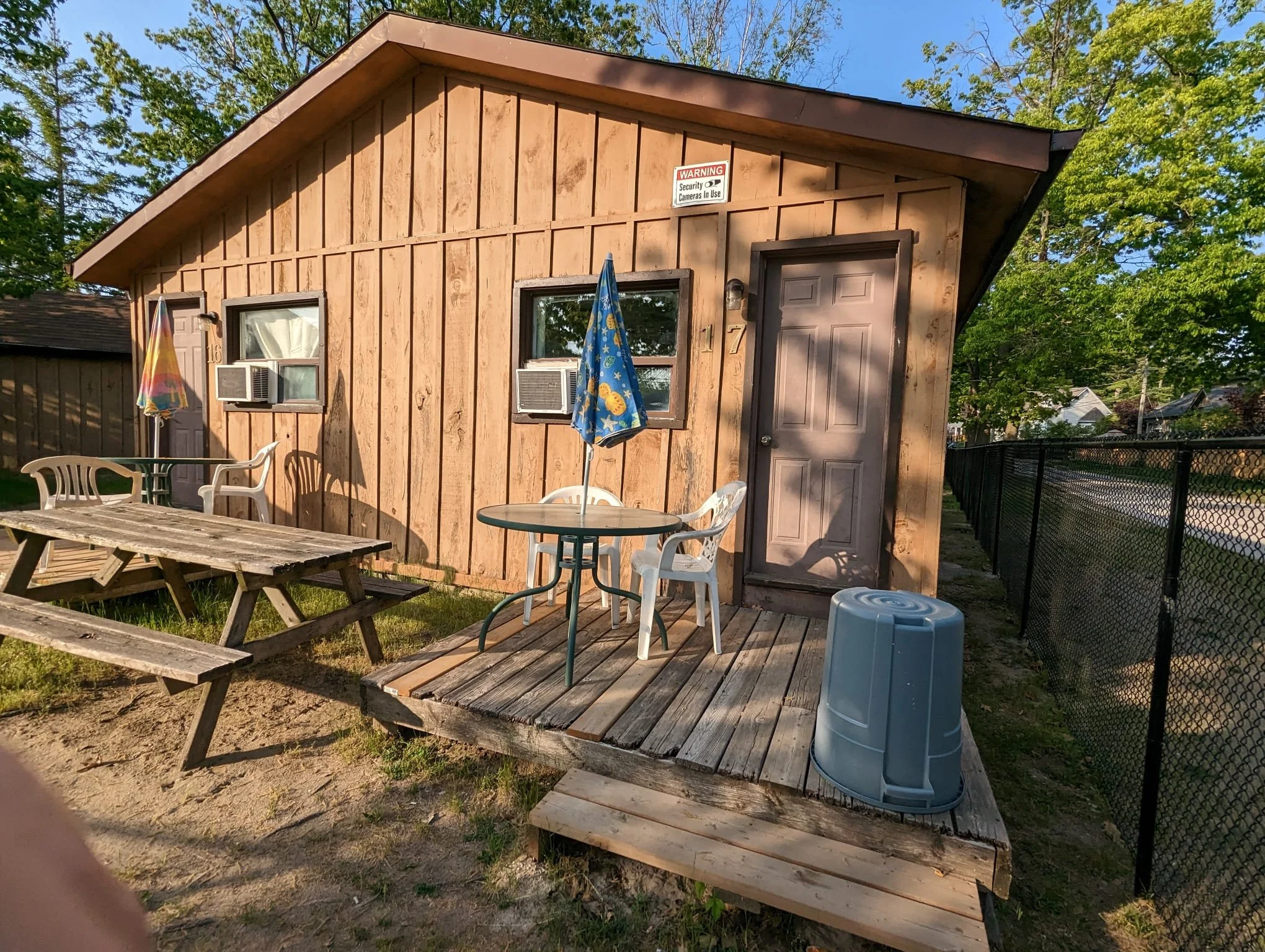 Backyard scene with small wooden building, two windows with air conditioning units, a gray door, and a small wooden deck. The deck has a round table with two chairs, umbrellas, a plastic picnic table, and a trash can. Green trees and a black fence are in the background.