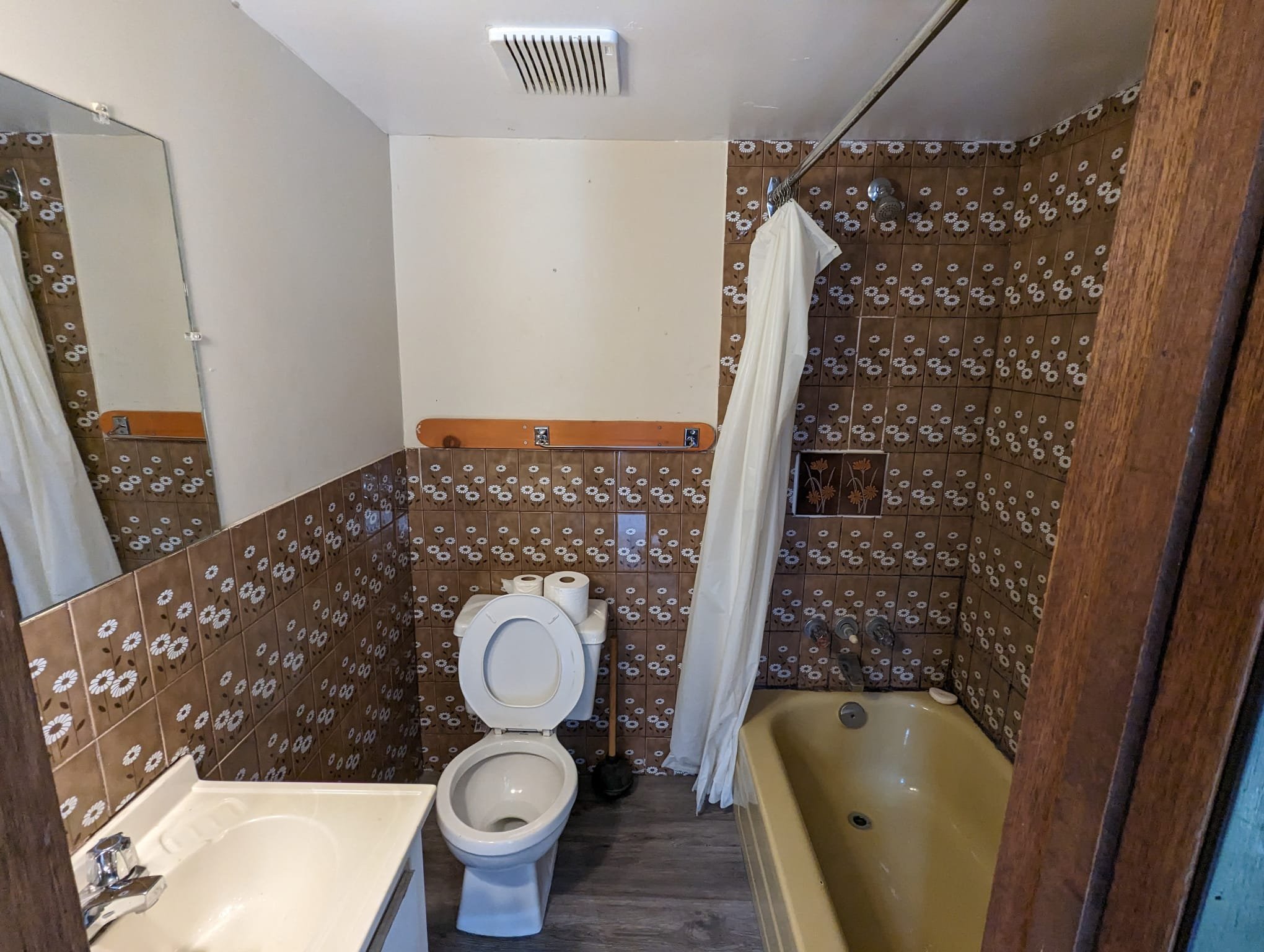 A small vintage bathroom with brown floral tiled walls, a yellow bathtub, a white toilet with rolls of toilet paper on top, a small white sink, a mirror, and a curtain rod with a white shower curtain. The floor is wood laminate, and the ceiling has a ventilation fan.