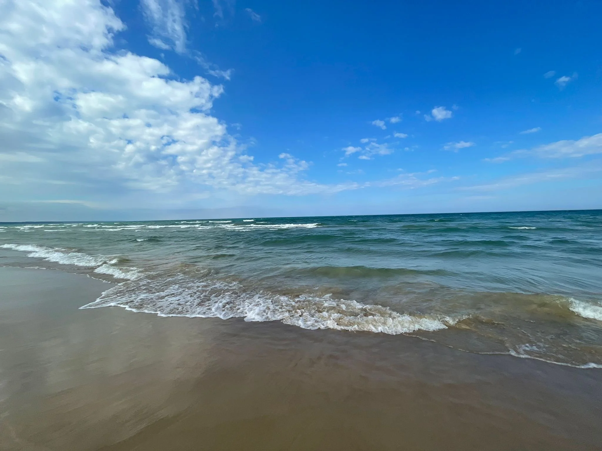 Atlantic Ocean with waves gently lapping a sandy beach under a partly cloudy blue sky.