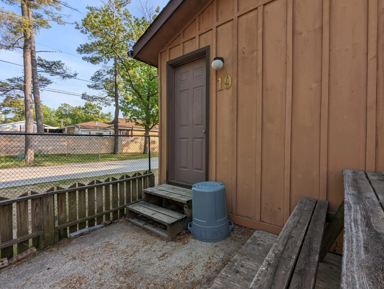 Front porch of a house with a closed door, house number 19, a light above the door, a blue trash can upside down, small wooden steps, and a wooden bench, surrounded by a chain-link fence, trees, and neighboring houses in the background.