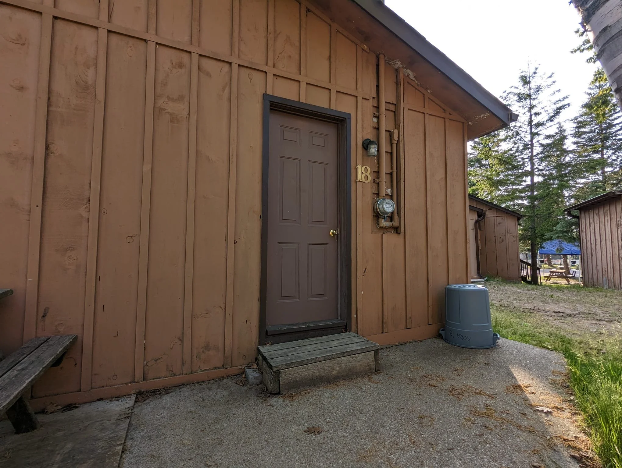 Brown wooden exterior of a house with a door, house number 18, electrical meter, and trash bin outside.