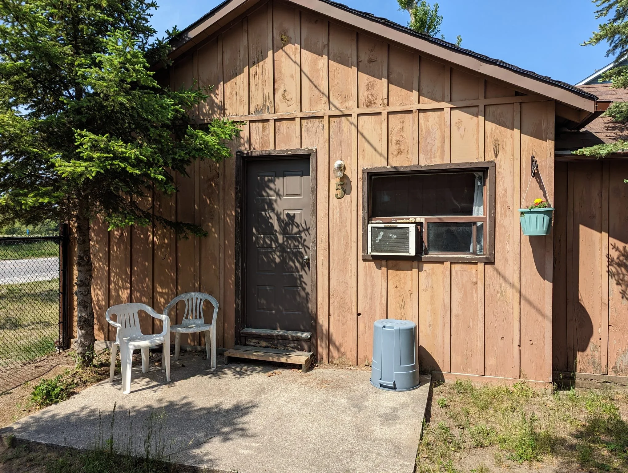 Exterior of a small wooden house with a brown door, small window with an air conditioning unit, a hanging planter, two white plastic chairs, and a gray trash bin on a concrete patio, surrounded by greenery and a black fence.
