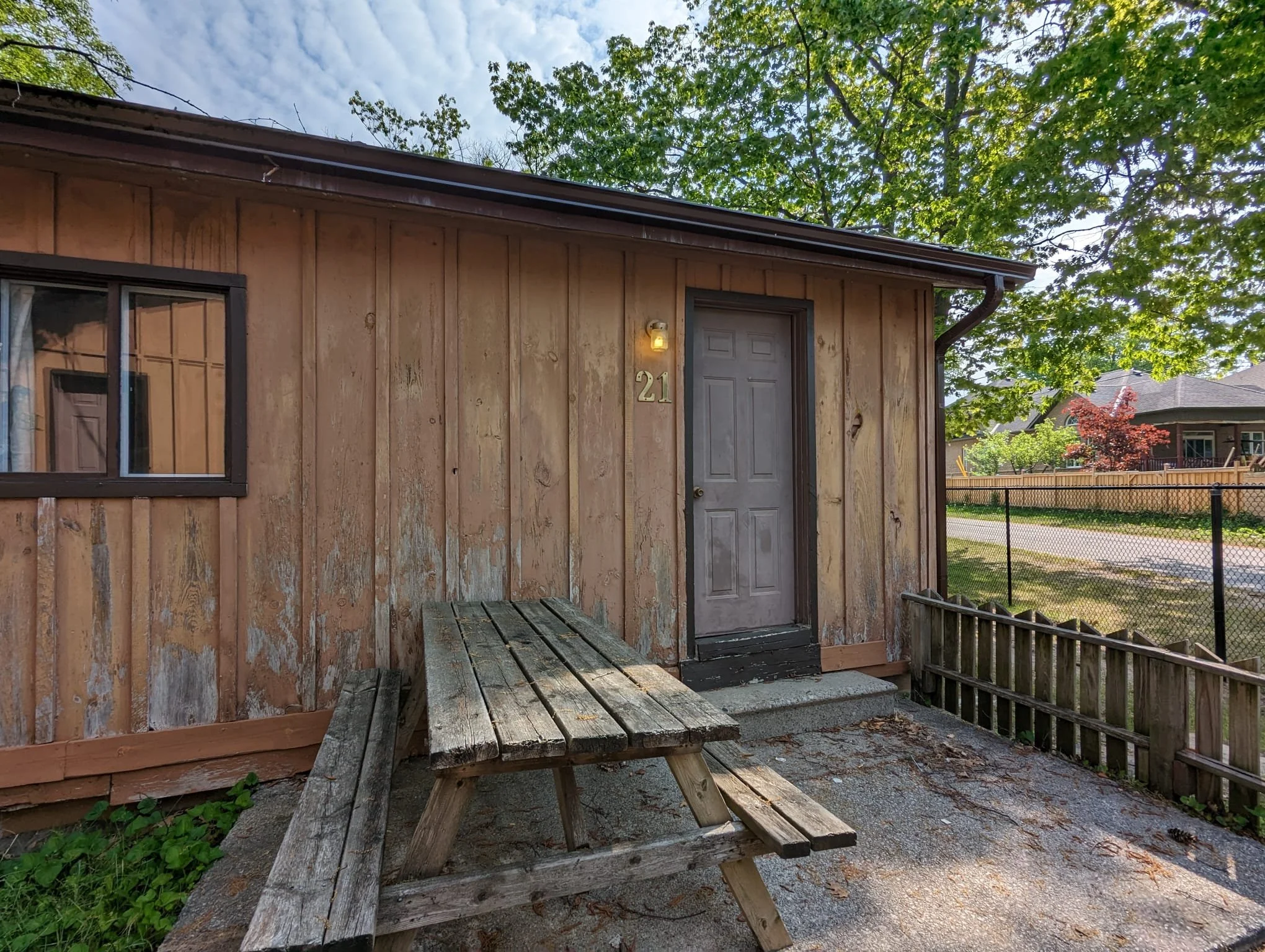 Front of a small house with weathered wooden siding, a grey door, a window on the left, and a wooden picnic table in front. The house number 21 is displayed next to the door. There are trees and neighboring houses in the background.