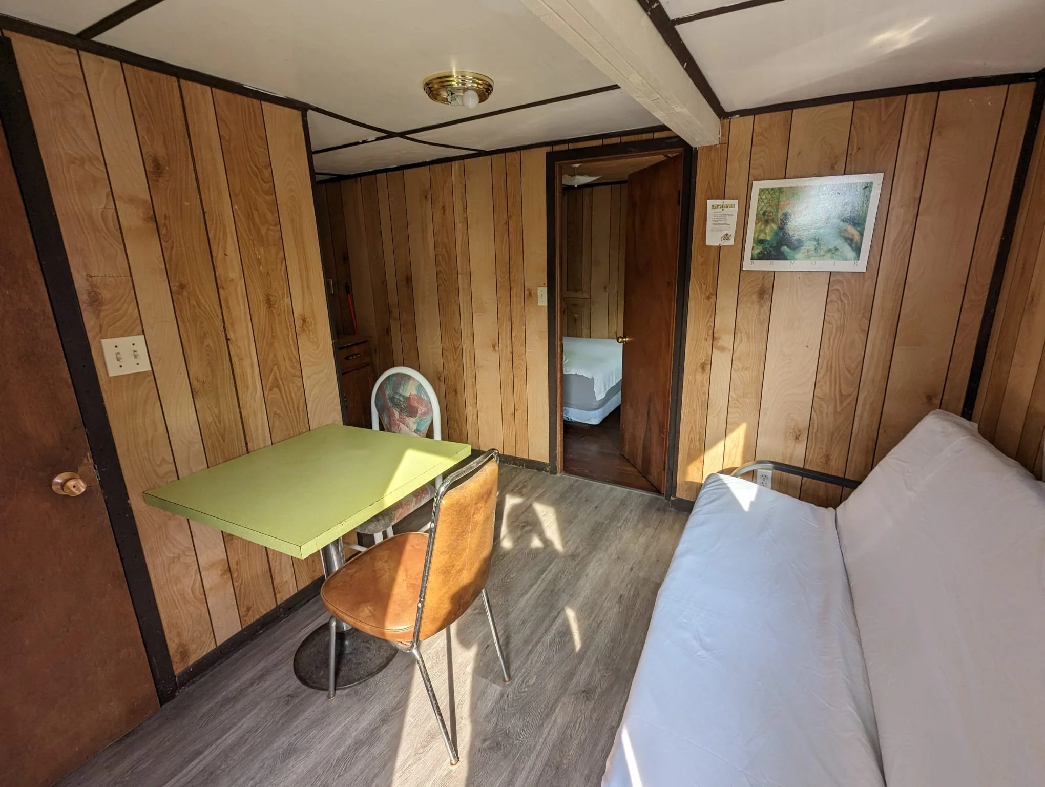 Small room with wood-paneled walls, a white bed, a green table, two chairs, and a framed landscape picture on the wall.