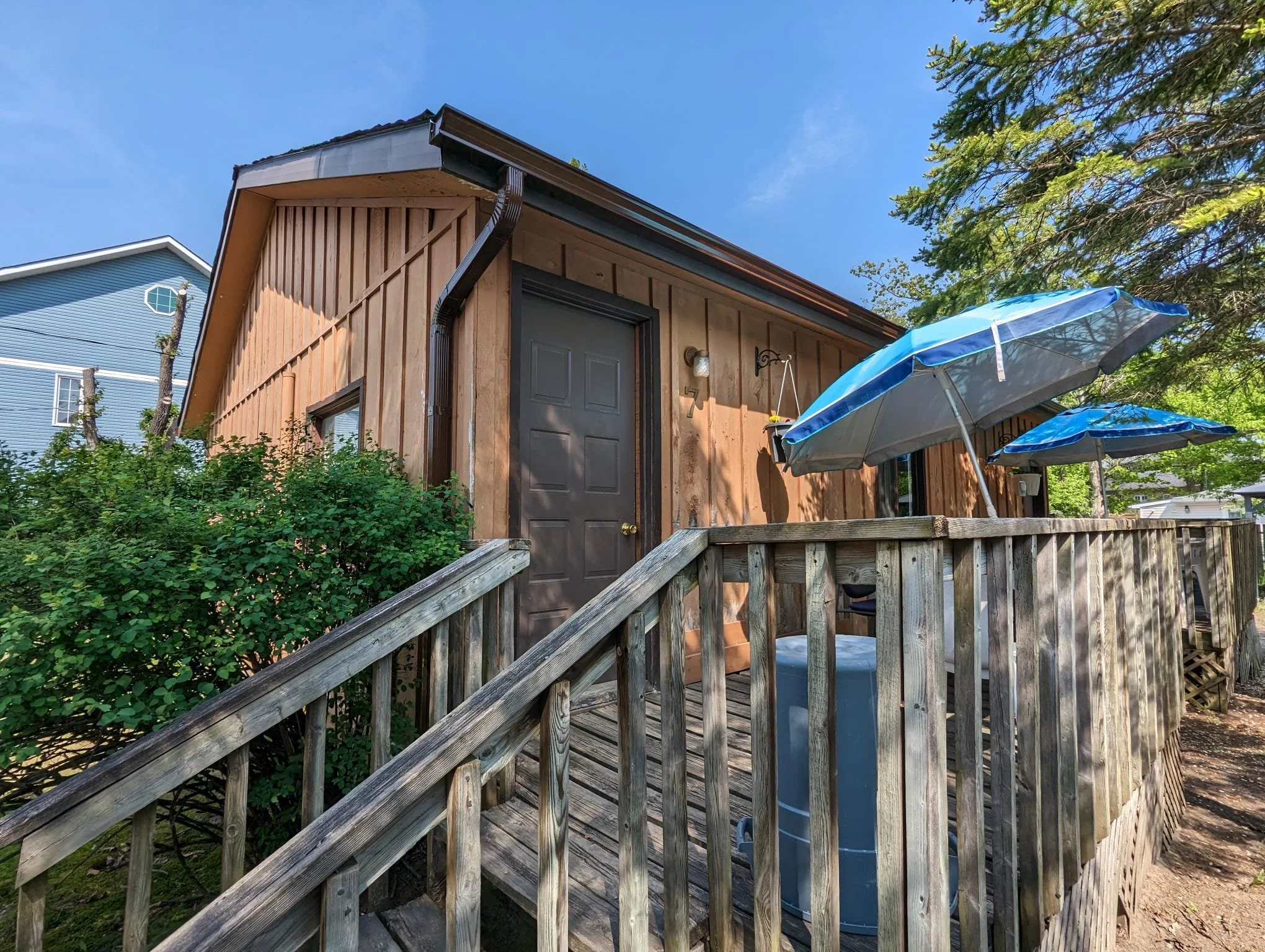 Wooden house with a deck, two blue umbrellas, and greenery on a sunny day.