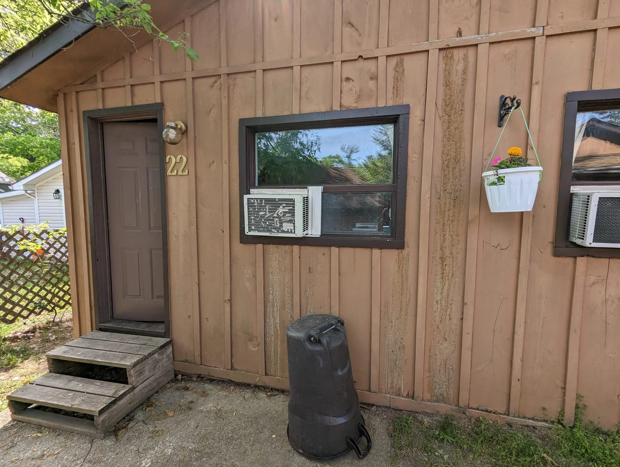 Front of a house with brown wooden siding, a door with the number 22, two windows with air conditioning units, a white hanging flower pot, and a black trash can on a concrete and dirt surface.