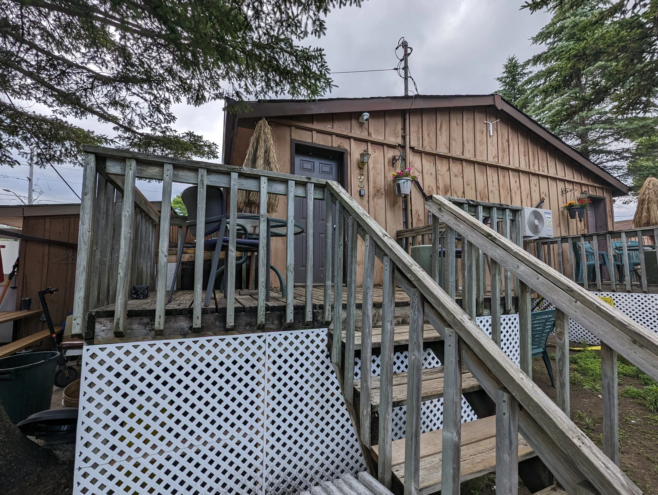 Wooden house with stairs leading up to a deck, outdoor furniture, potted plants, and a large tree overhead.