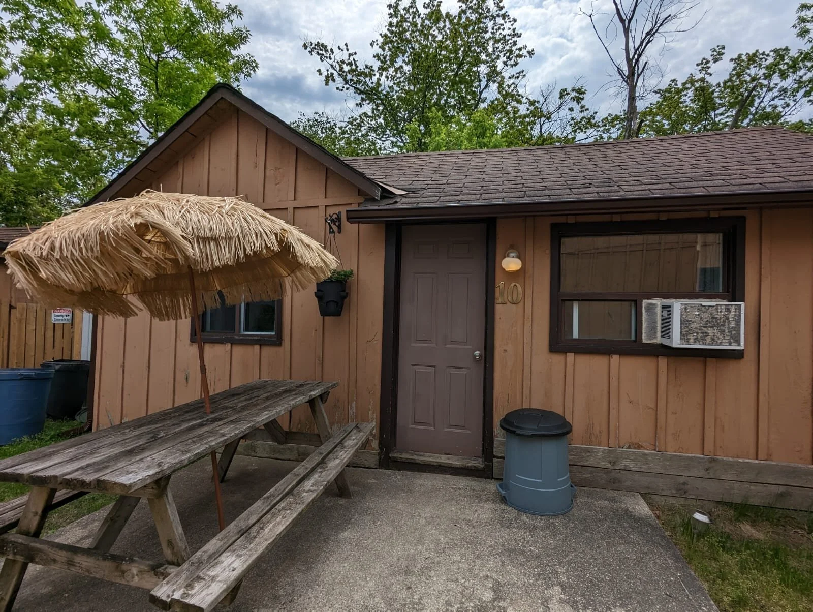 Front of a small house with a brown door, window with an air conditioning unit, and a wooden picnic table with a straw umbrella outside.