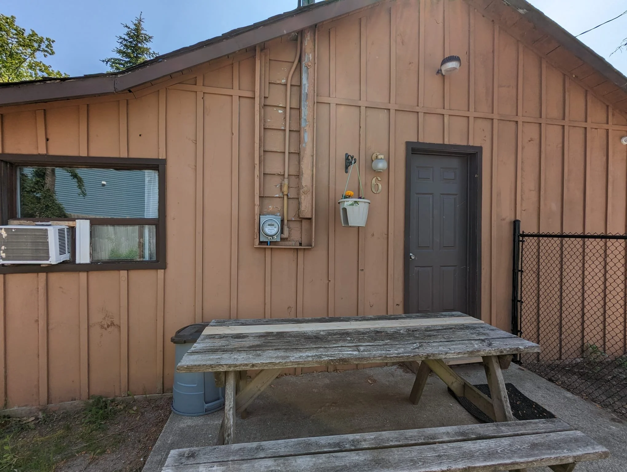 The exterior of a small wooden building with a dark gray door, a window with an air conditioning unit, and a hanging flower planter. There is a wooden picnic table in front of the building and a chain-link fence on the right side.