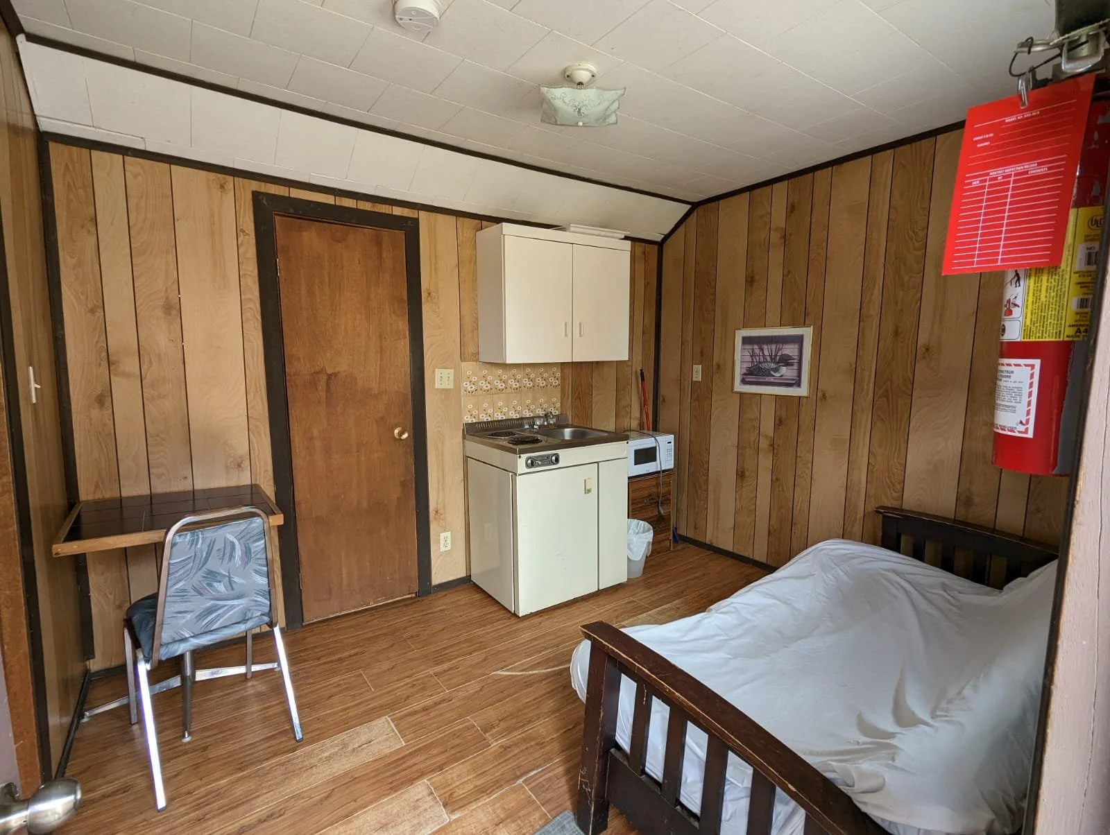 Small room with wood-paneled walls, a single bed with a white sheet, a mini kitchenette including a small stove, microwave, and cabinet, a wooden table with a metal chair, a framed picture on the wall, and a fire extinguisher on the wall.