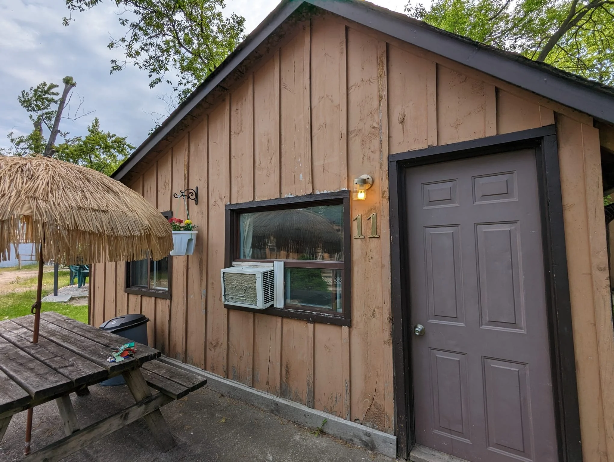 Small wooden building with a dark gray door and two windows, one with an air conditioning unit, labeled with the number 11. There is a picnic table with a thatched umbrella and some hanging flower pots outside.