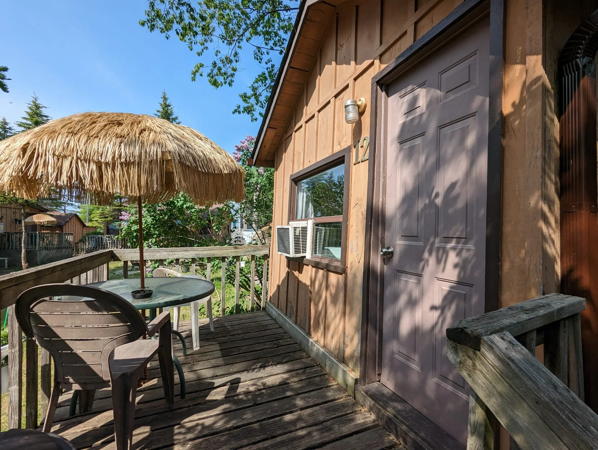 Wooden balcony with a plastic chair, a table with a thatched umbrellas, and an exterior door of a wooden house with a window and an air conditioning unit. Trees and other houses are visible in the background.