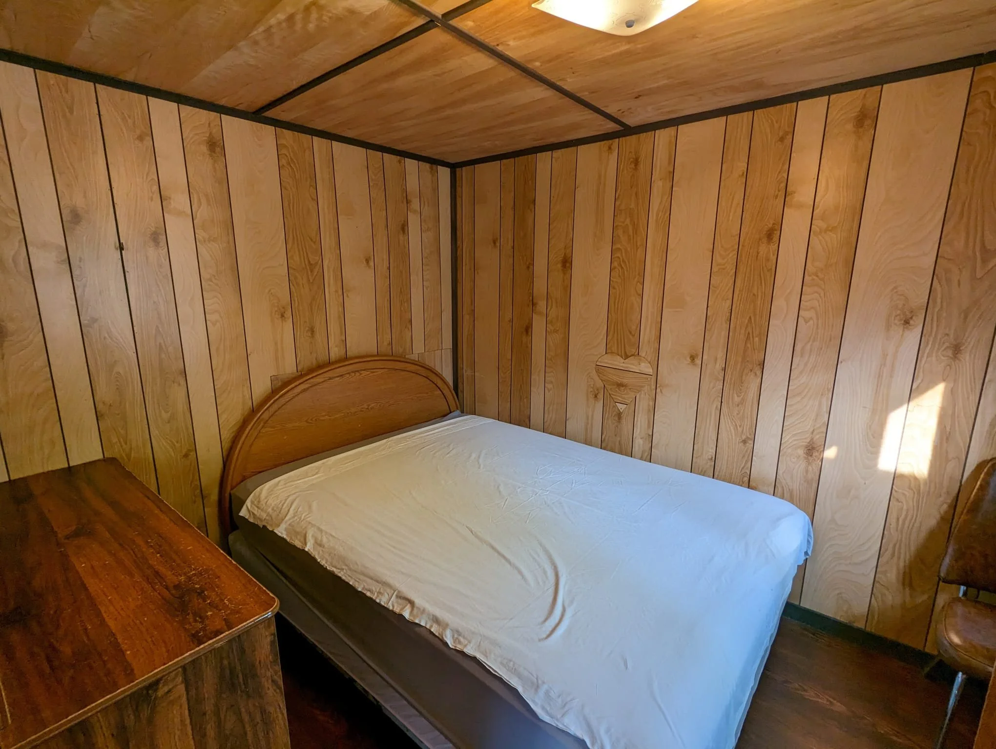 A wooden-paneled bedroom with a bed, nightstand, and chair, illuminated by ceiling light and sunlight through a window.