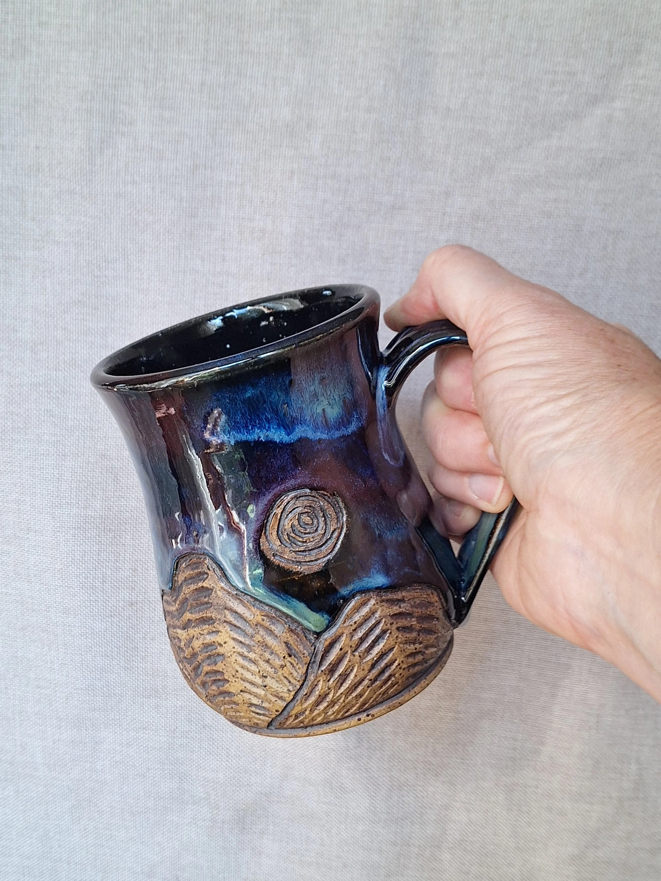 A ceramic mug with a glossy black and blue glaze on the upper part and a carved earthy brown mountain design on the lower part, held by a person's hand against a plain light-colored background.