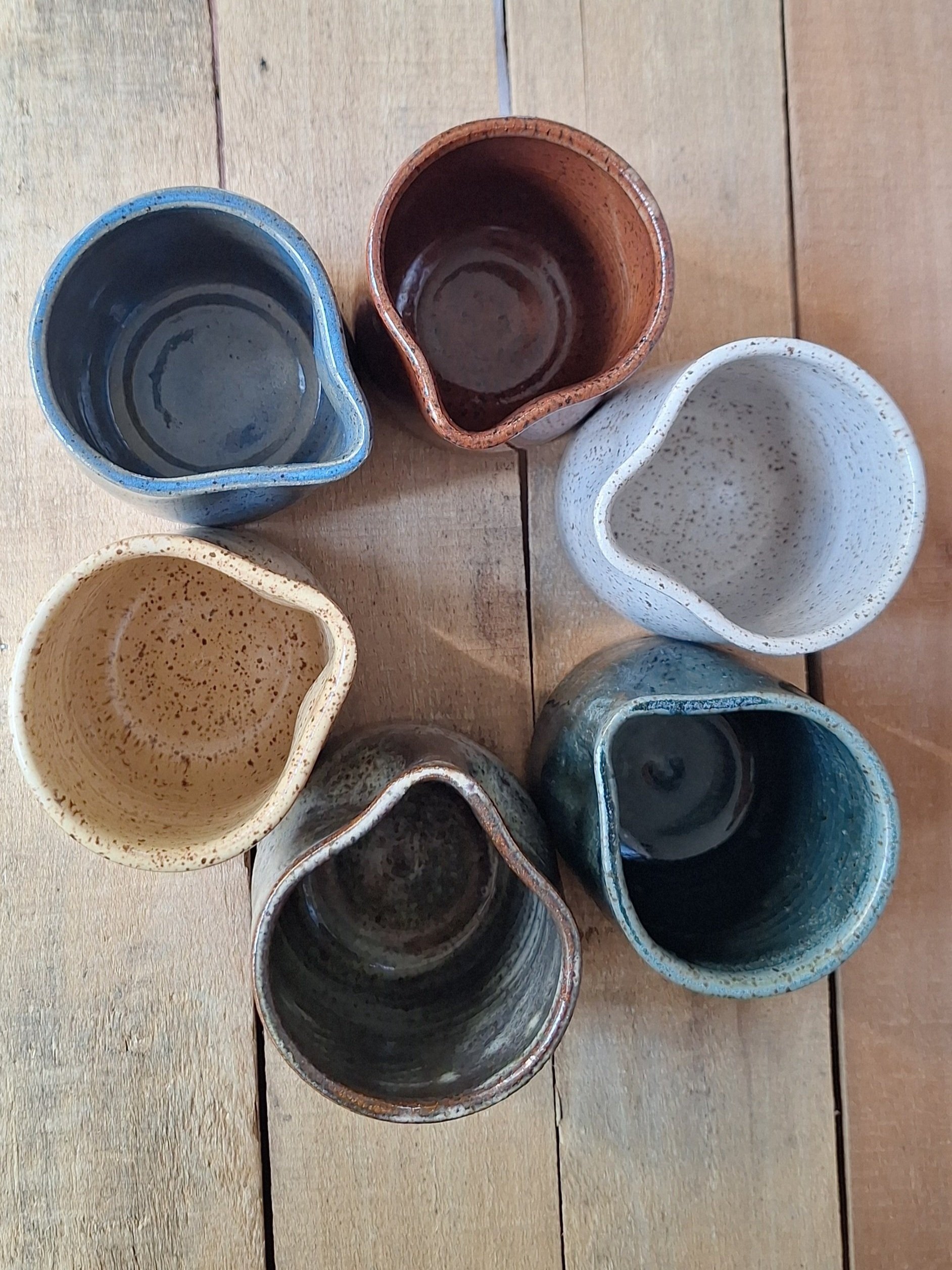 Six small ceramic sauce cups arranged in a circle on a wooden surface, each with different glaze colors and speckled patterns.