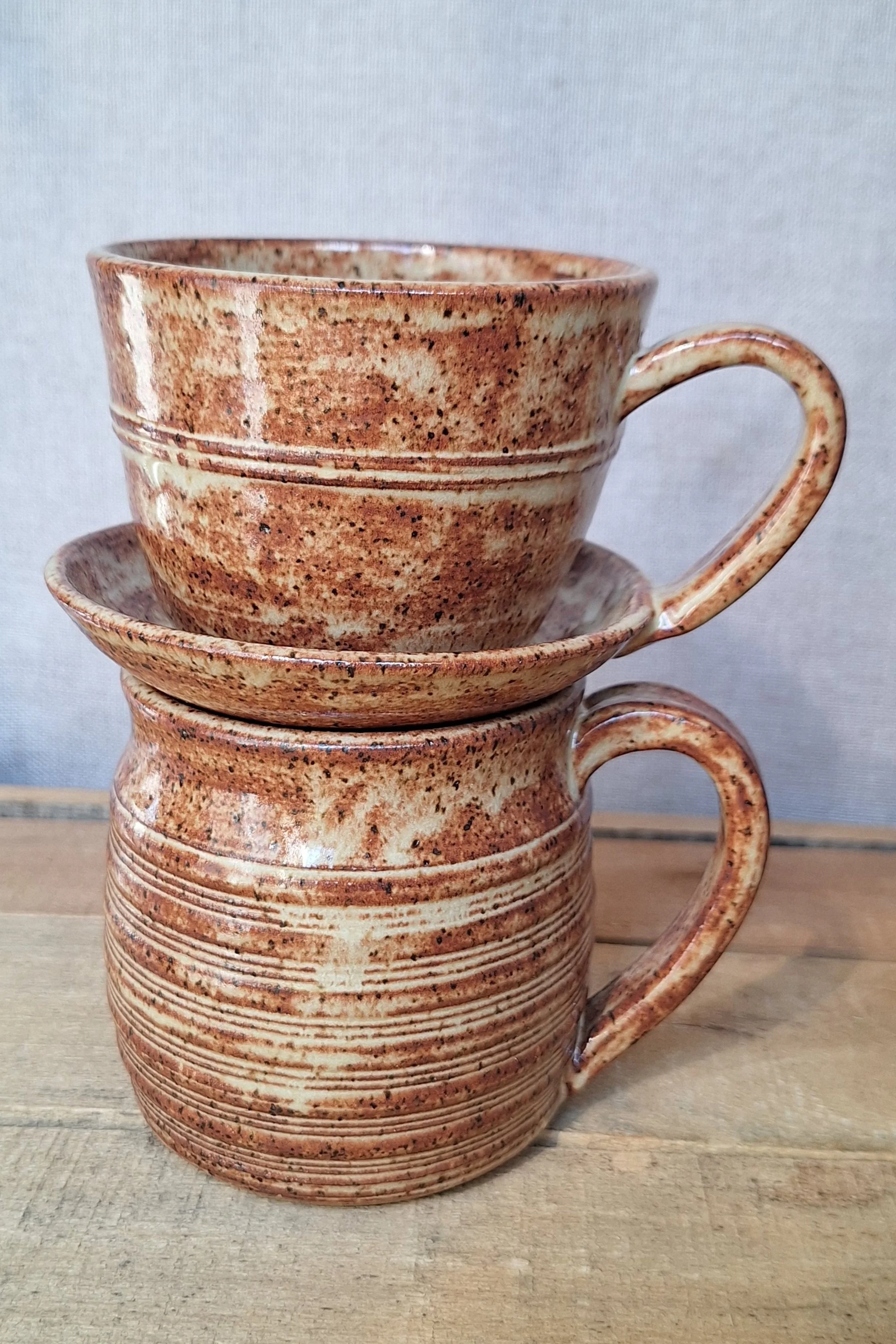 Two stacked ceramic mugs with handles, featuring a speckled brown and beige glaze, sitting on a wooden surface against a gray background.